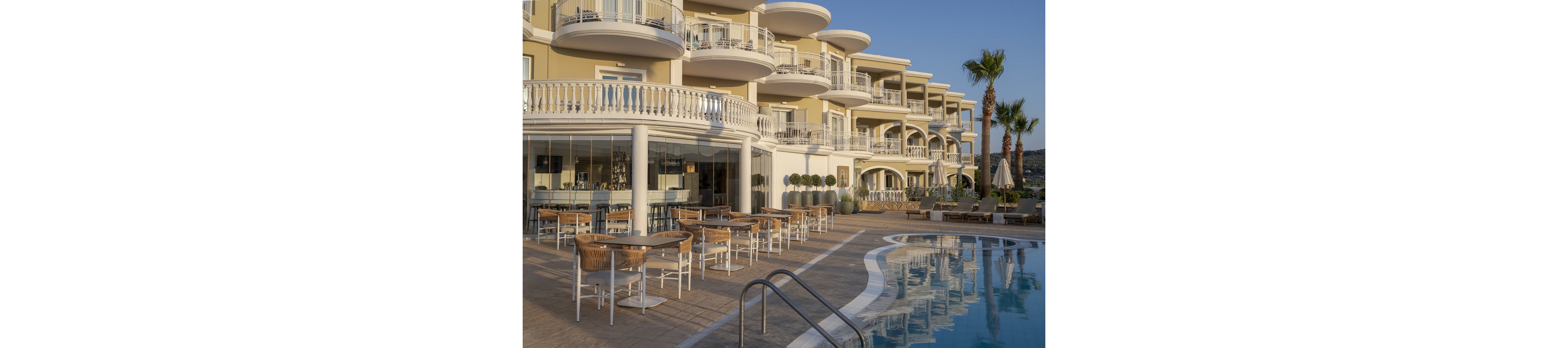 A modern luxury hotel features elegant balconies and a spacious outdoor pool area. Wicker chairs and tables are arranged near the pool, inviting guests to relax in the warm evening light. The architecture is characterized by soft beige tones and curved lines, complemented by palm trees and manicured landscaping. The tranquil setting suggests a resort atmosphere, ideal for leisure and relaxation.