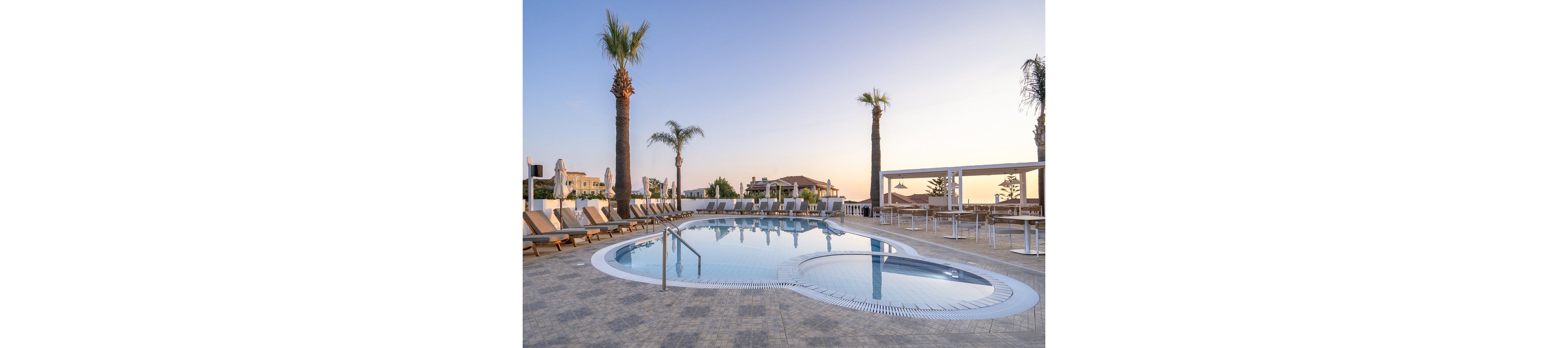 A tranquil outdoor swimming pool is surrounded by lounge chairs and tall palm trees, set in a modern resort environment. The scene is captured at sunset, with soft pastel colors in the sky and gentle reflections on the water. The area is clean and well-maintained, with no people present, evoking a calm and relaxing mood. Buildings and shaded seating areas are visible in the background, enhancing the vacation atmosphere.
