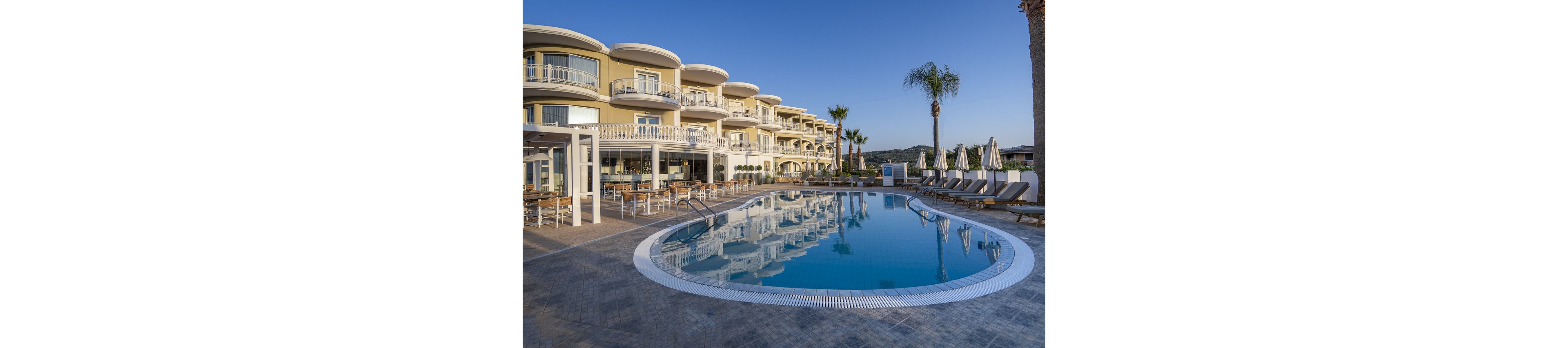 A contemporary multi-story hotel features curved balconies and overlooks a large, clear outdoor swimming pool. Sun loungers and umbrellas are neatly arranged around the poolside, with palm trees adding a tropical touch. The setting is calm and inviting, with clear blue skies and soft natural lighting enhancing the relaxed vacation atmosphere.