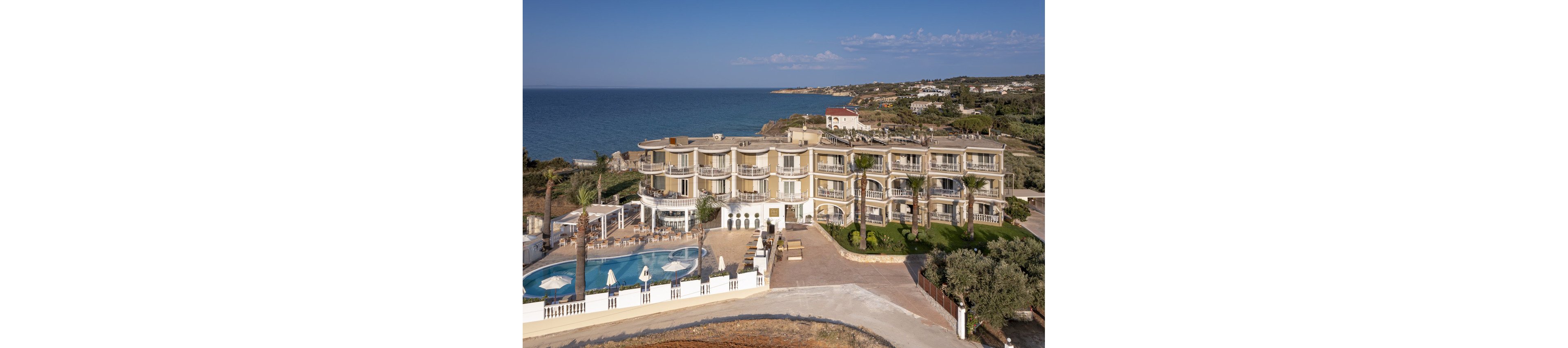 A multi-story hotel building is situated along a scenic coastline, offering expansive views of the ocean. The property features a large outdoor swimming pool surrounded by lounge chairs and palm trees, creating a relaxing atmosphere. The architecture is modern with curved balconies, and the setting is tranquil with clear skies and lush greenery nearby. No visible text or numbers are present in the image.
