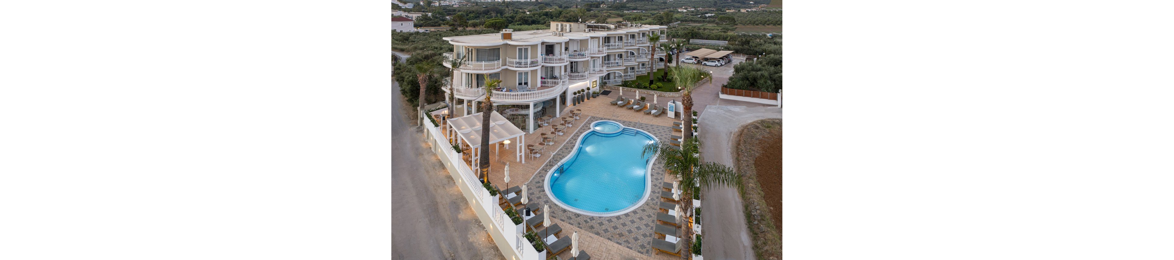 This image shows a contemporary multi-story hotel featuring a large, uniquely shaped outdoor swimming pool. The pool area is surrounded by sun loungers, umbrellas, and palm trees, creating a relaxing atmosphere. The hotel is set in a semi-rural landscape with open fields and greenery in the background. The overall mood is tranquil and inviting, ideal for vacation or leisure stays.