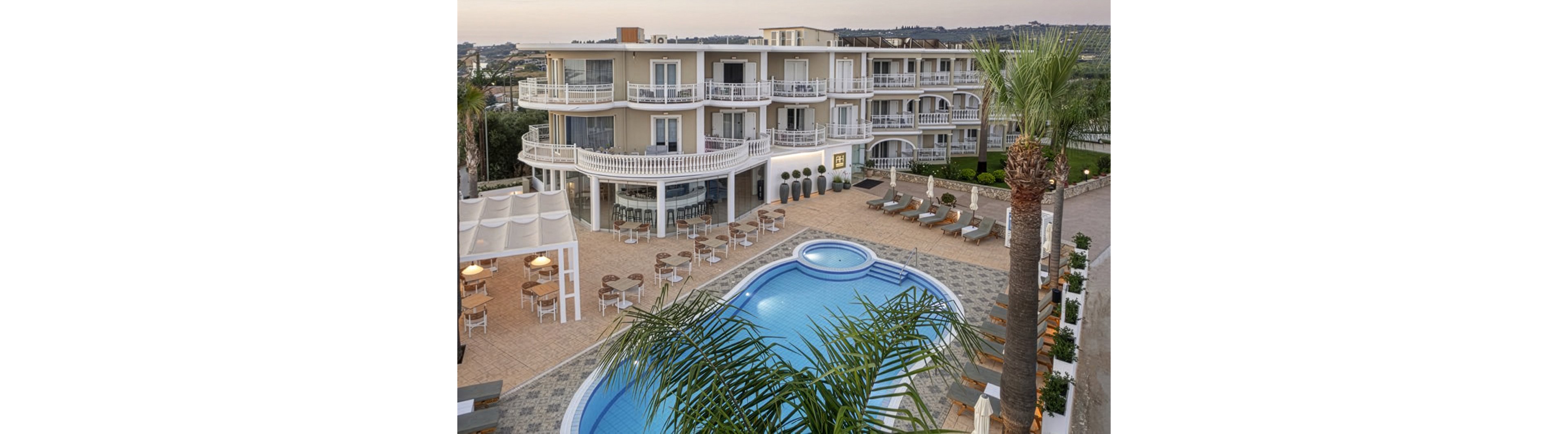 A multi-story resort hotel features balconies overlooking a curved outdoor swimming pool. Sun loungers and umbrellas are arranged neatly around the poolside terrace, with a shaded seating area and café tables nearby. Palm trees and landscaped greenery frame the scene, suggesting a warm coastal or Mediterranean destination. The overall atmosphere is calm and inviting, ideal for vacation relaxation.