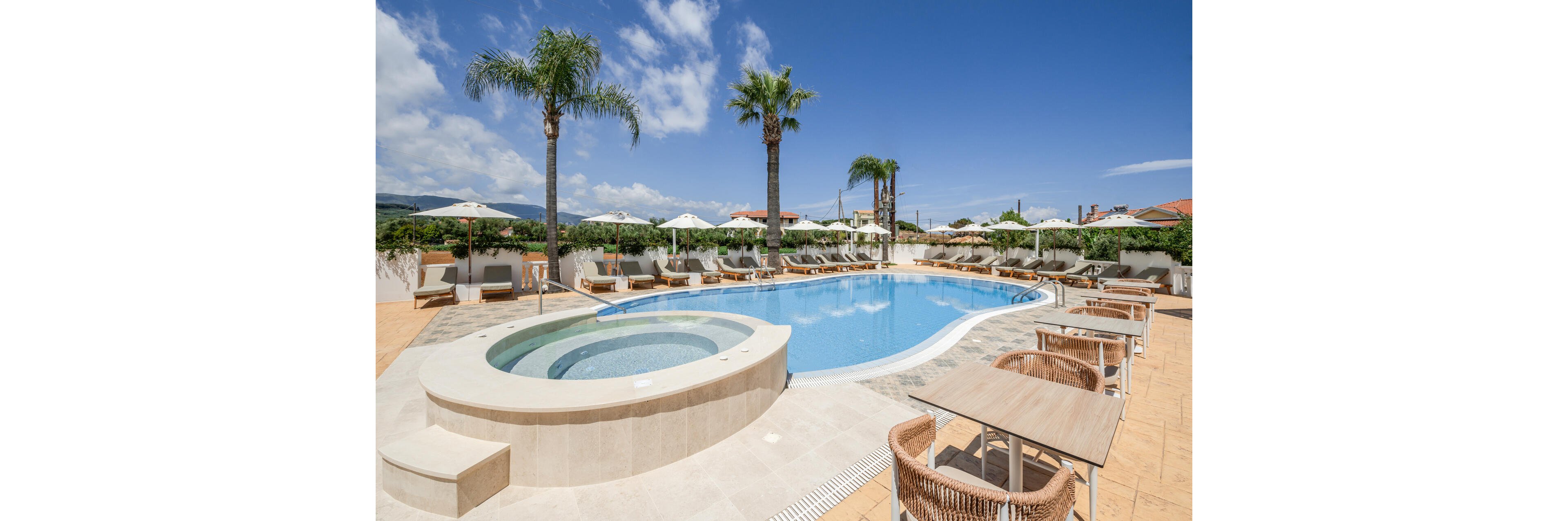 An inviting resort swimming pool is surrounded by lounge chairs and umbrellas under a bright blue sky. A circular hot tub or shallow pool is positioned in the foreground, leading into the larger curved main pool. Palm trees rise above the pool area, adding a tropical feel to the scene. Outdoor dining tables and woven chairs sit nearby on the tiled terrace, suggesting a relaxed vacation atmosphere.