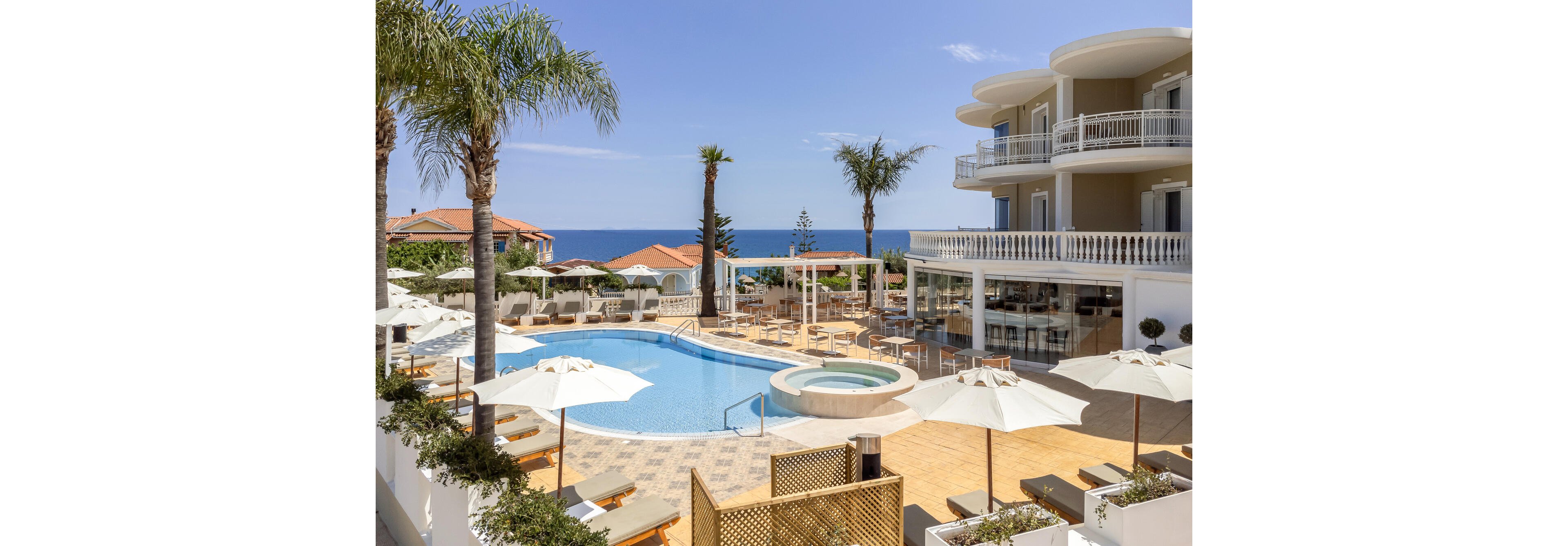 Upscale hotel with a curved outdoor swimming pool overlooks a calm blue sea. Sun loungers and white umbrellas surround the poolside terrace, creating a relaxing resort atmosphere. A circular hot tub and open-air bar area add to the leisure amenities. Modern multi-story buildings with balconies and palm trees frame the bright, sunny coastal scene.