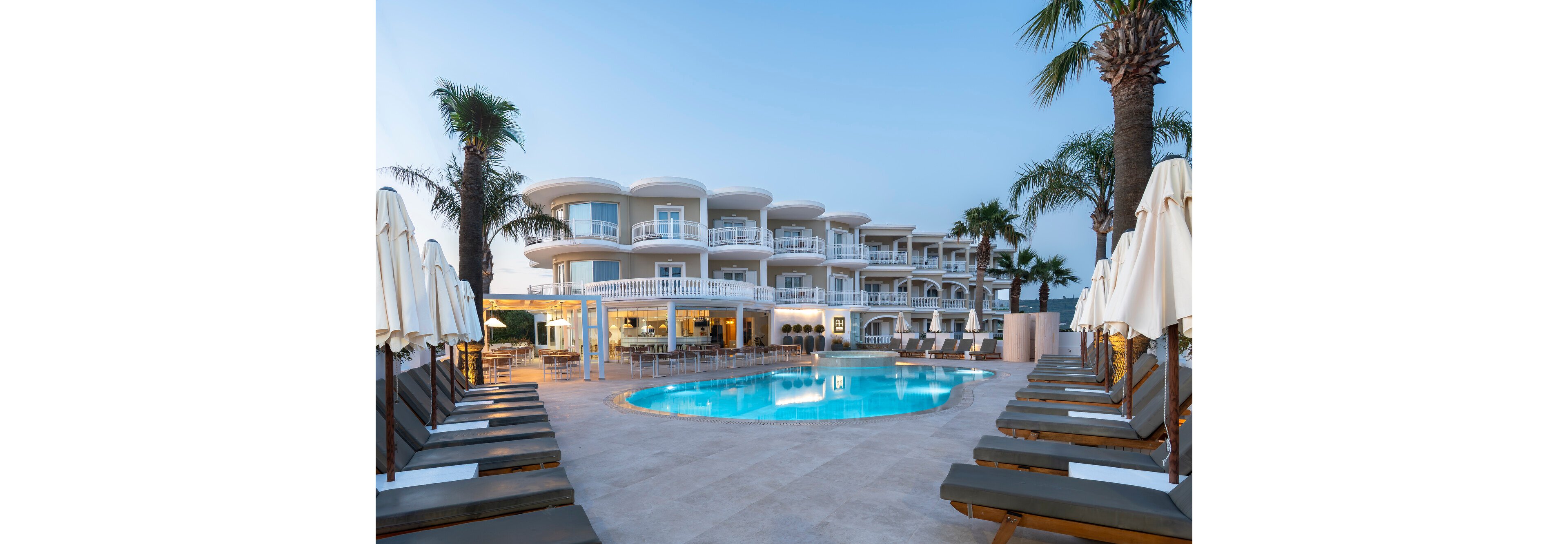 A contemporary multi-story hotel surrounds a curved outdoor swimming pool at dusk. Sun loungers and closed umbrellas line the poolside terrace, creating a symmetrical and inviting layout. Warm interior lights glow from the hotel rooms and restaurant area, contrasting with the cool blue water and evening sky. Palm trees frame the scene, enhancing the relaxed, resort-style atmosphere.