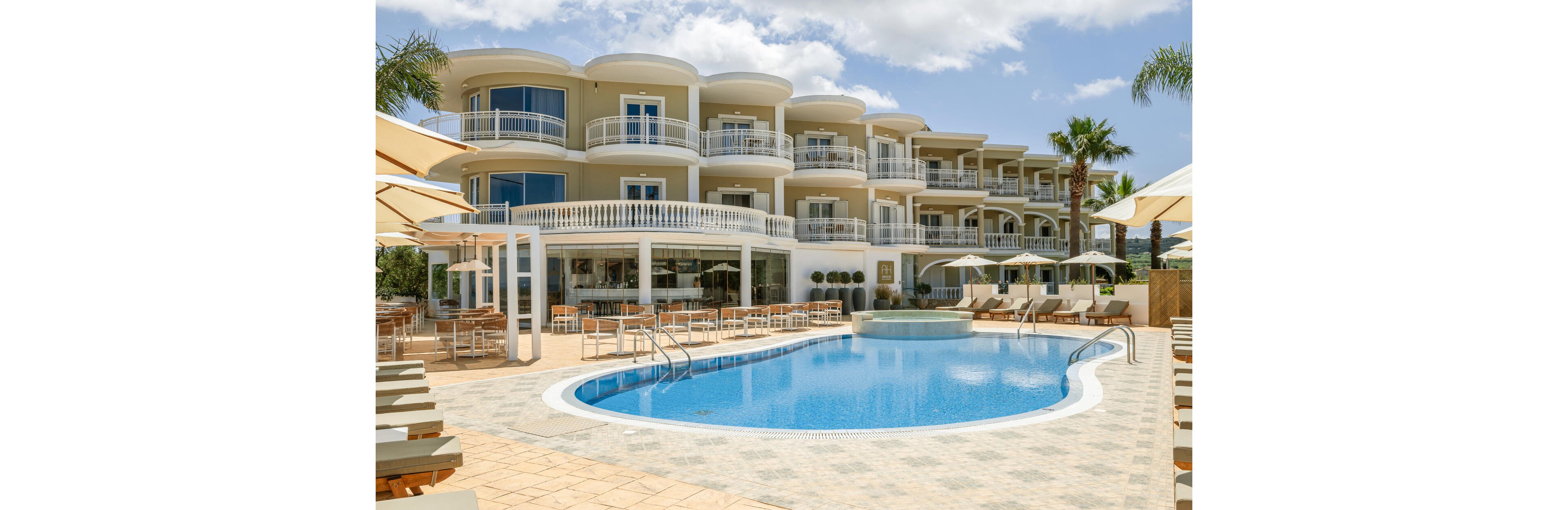 A contemporary multi-story resort hotel features curved balconies overlooking a clear blue outdoor swimming pool. Sun loungers and umbrellas surround the poolside terrace, creating a relaxing vacation atmosphere. Palm trees and neatly arranged outdoor seating enhance the tropical, sunny setting. The sky is bright with scattered clouds, suggesting a warm and inviting holiday destination.
