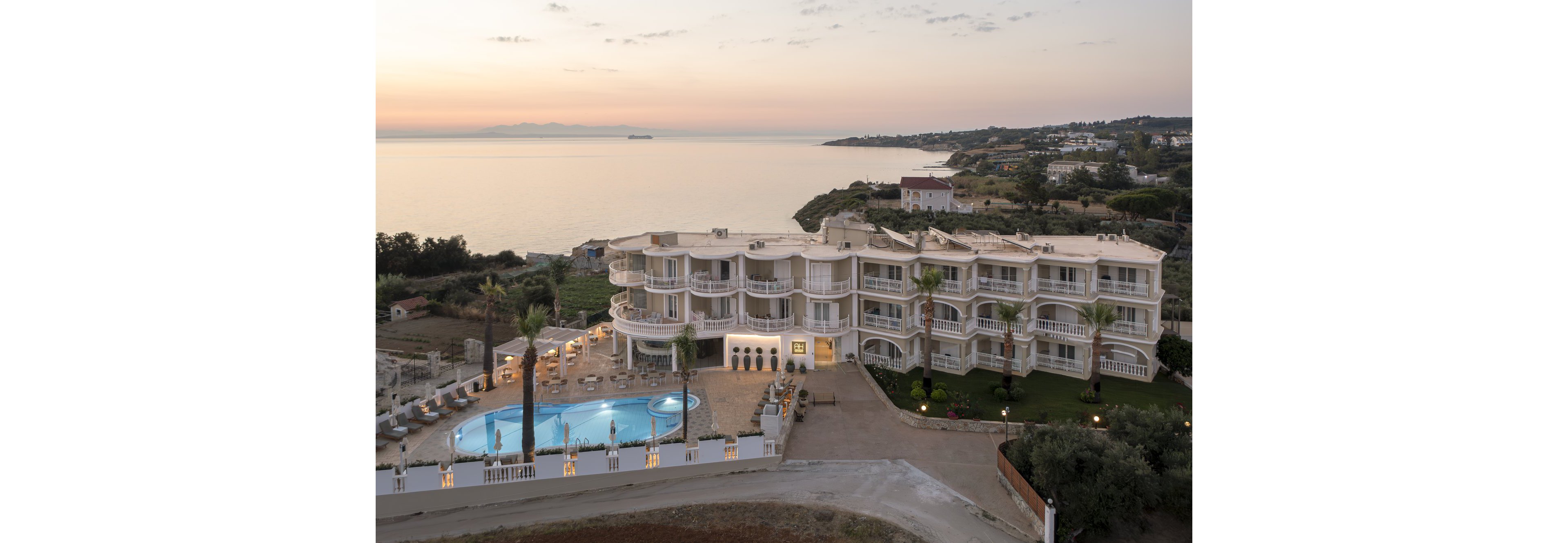 A large seaside resort hotel is shown from an elevated viewpoint at sunset. The multi-story building features numerous balconies facing the calm sea and coastline. A bright blue outdoor swimming pool with surrounding sun loungers stands in front of the hotel. The scene includes palm trees, nearby houses, and a distant ship on the tranquil water under a pastel sky.