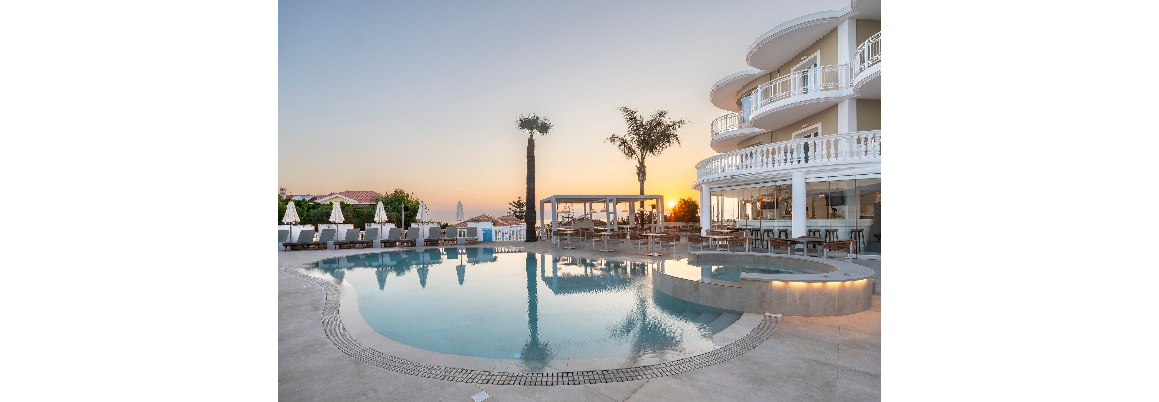 A curved infinity-style swimming pool sits beside a modern seaside hotel at sunset. White balconies and glass railings overlook the water, with sun loungers and umbrellas arranged neatly along the poolside. A few palm trees and a covered outdoor dining area enhance the resort atmosphere. The warm evening light reflects on the calm pool surface, creating a tranquil vacation setting.