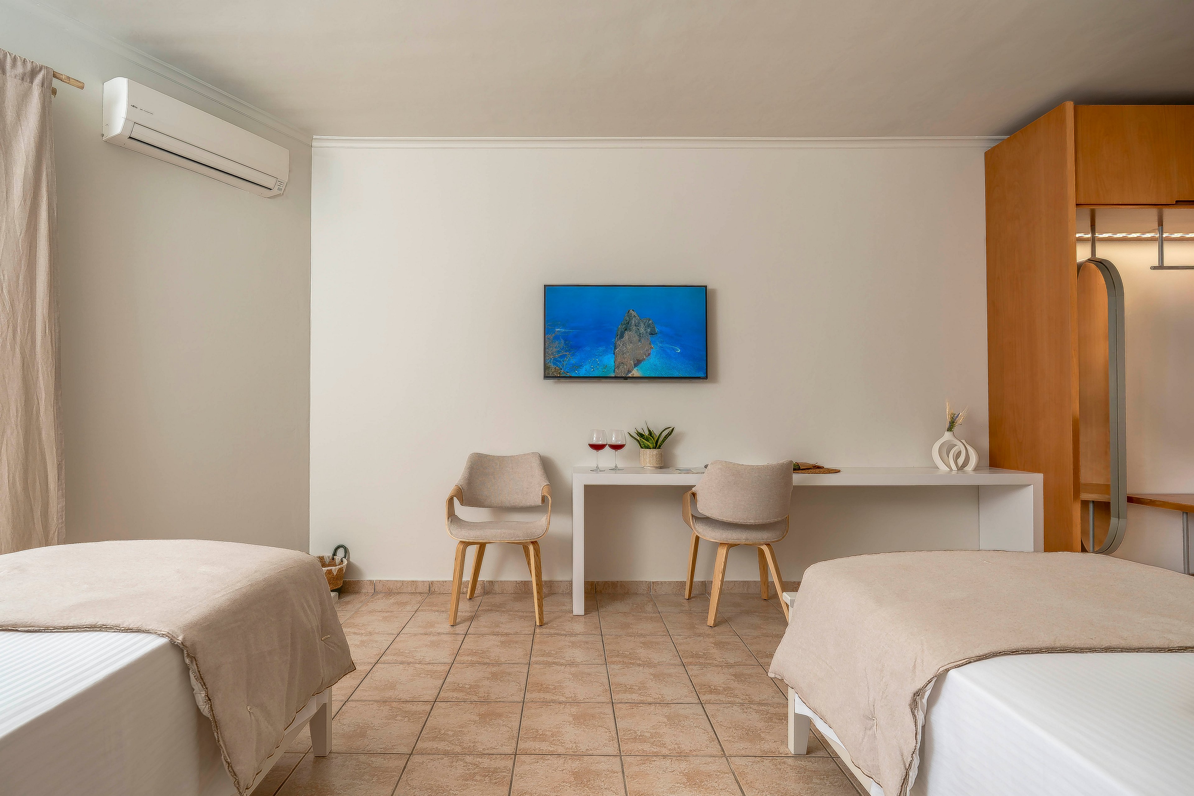 A serene spa treatment room features two neatly made massage beds facing a simple white wall. At the center, a flat-screen television displays an underwater scene above a long white desk with two modern wooden chairs. Soft beige linens, tiled flooring, and neutral curtains create a calm, minimalist atmosphere. A small plant and sculptural decor add subtle accents, while an air conditioning unit suggests a comfortable, climate-controlled space.