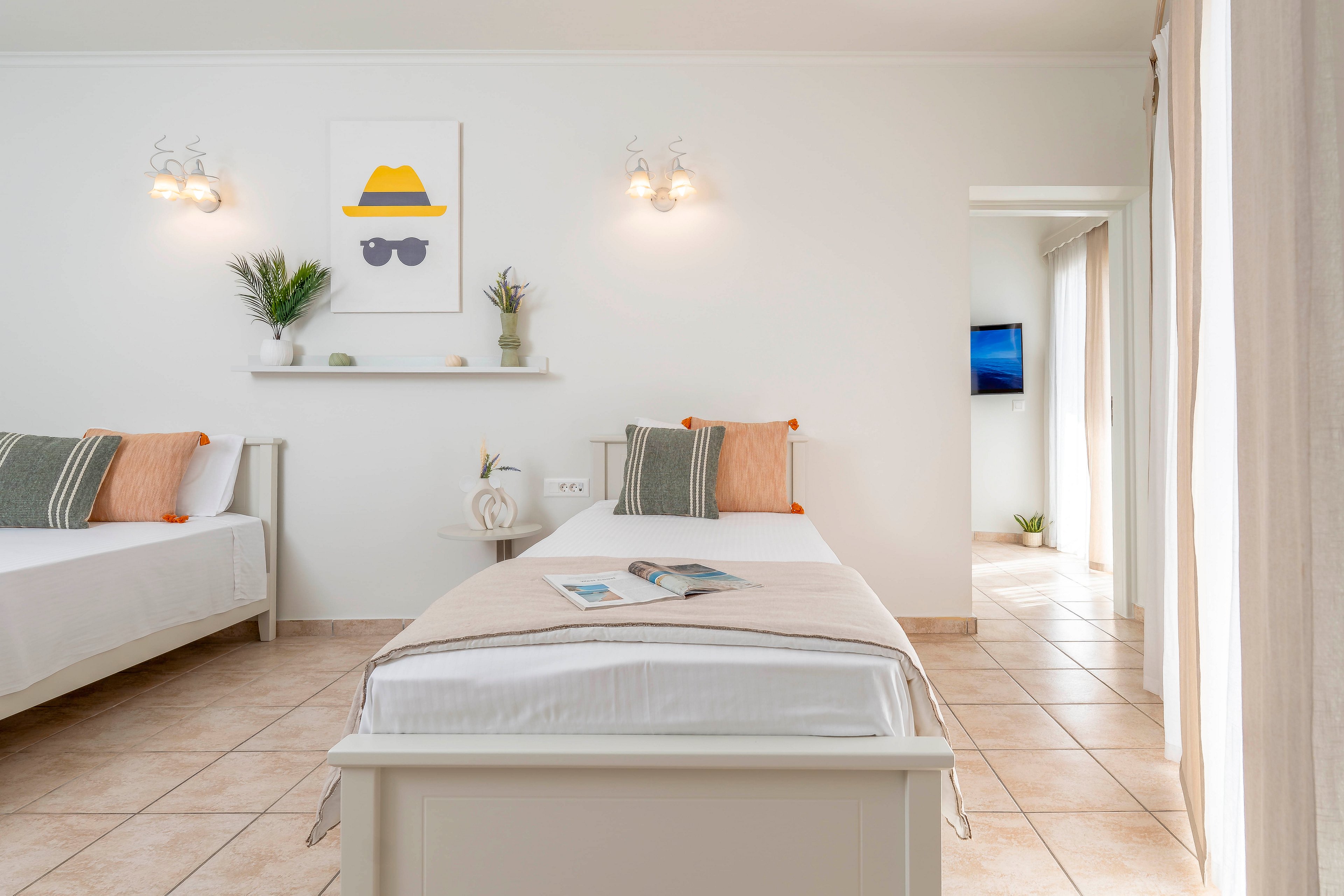 A bright, minimalist bedroom features two single beds with neutral bedding and soft accent pillows. The room has light tiled floors, white walls, and simple wall-mounted lights above a decorative shelf. A framed artwork with a yellow hat and sunglasses adds a playful touch to the otherwise calm decor. In the background, a doorway opens to a corridor with natural light and a wall-mounted television.