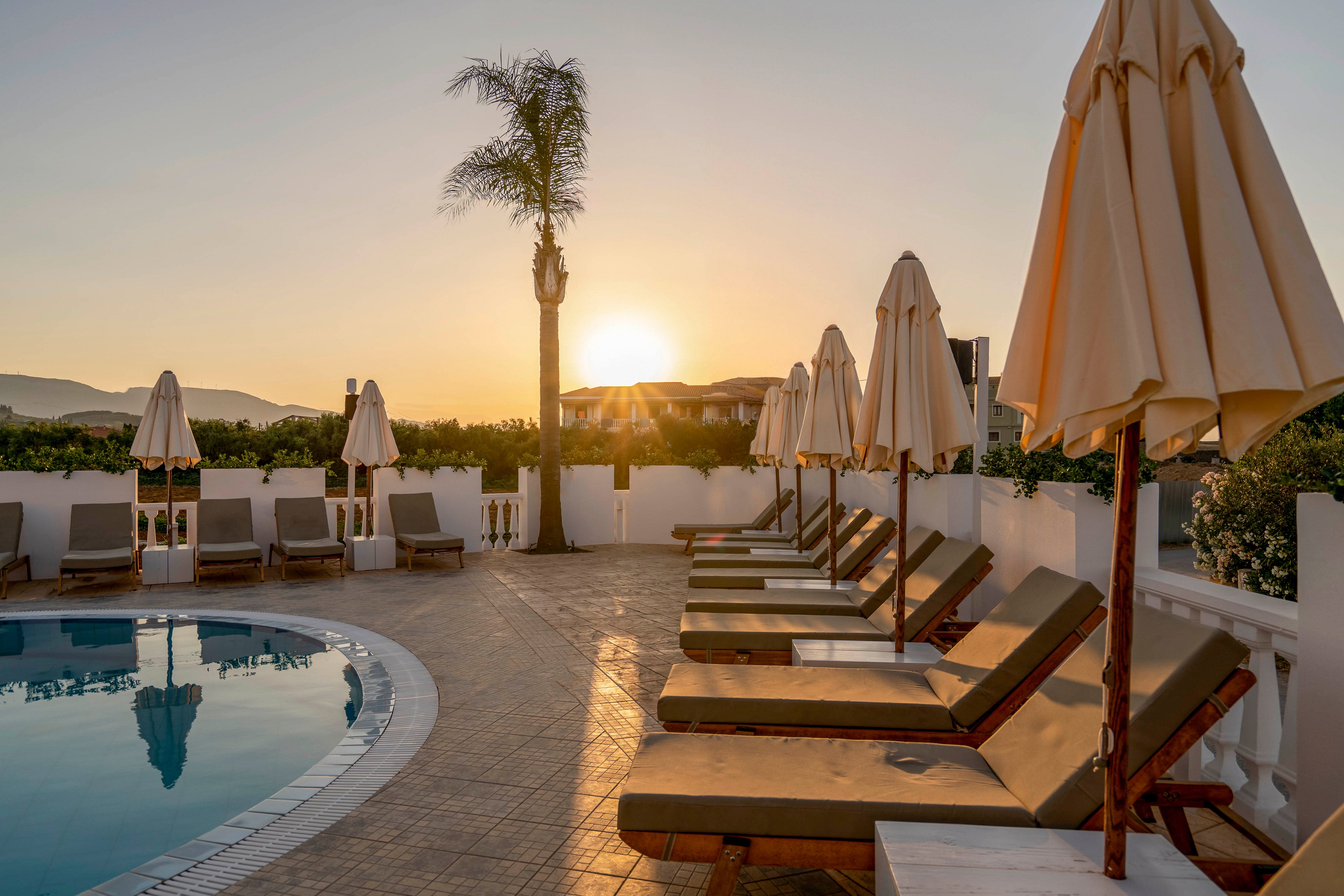A serene resort pool area is captured at sunrise with the sun low on the horizon. Wooden sun loungers with cushions line the tiled deck, each paired with a closed umbrella. The curved swimming pool reflects the warm morning light, while white walls and distant hills frame the scene. The atmosphere feels calm and inviting, suggesting a peaceful vacation setting.