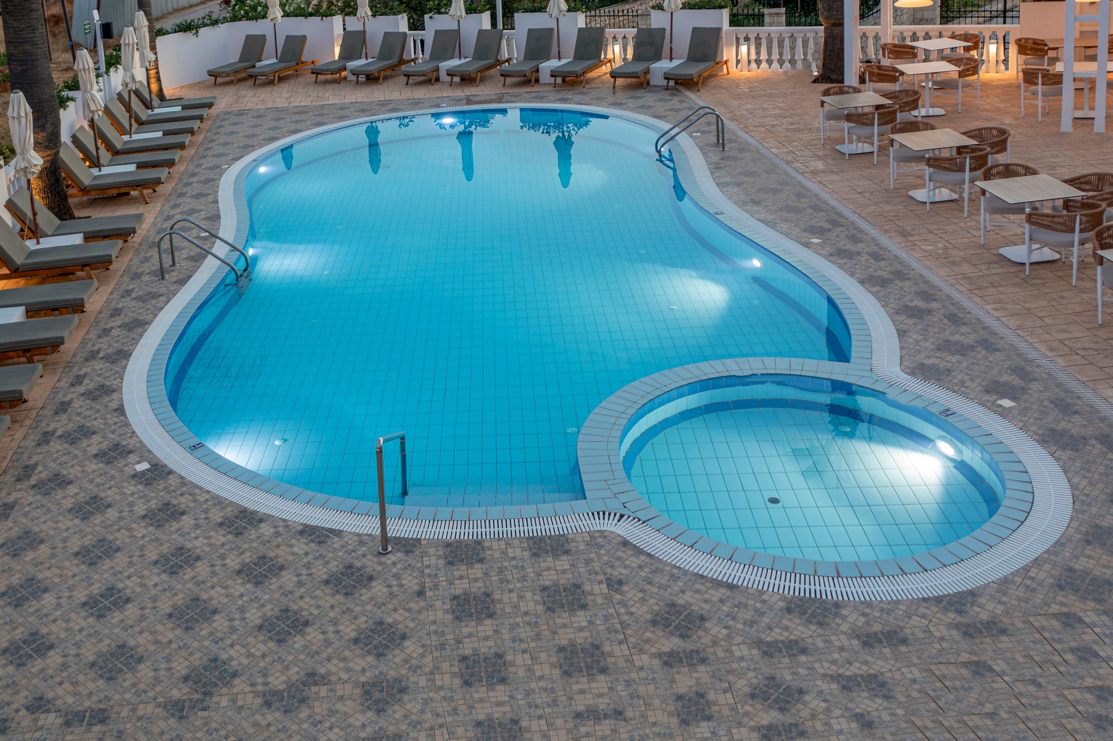 A clean, curving swimming pool with an attached circular kiddie pool sits in the center of a tiled terrace. Lounge chairs with cushions line the sides, each paired with small tables and umbrellas. In the background, additional seating and white railings suggest a hotel or resort setting. Soft evening light reflects on the calm blue water, creating a tranquil and inviting atmosphere.