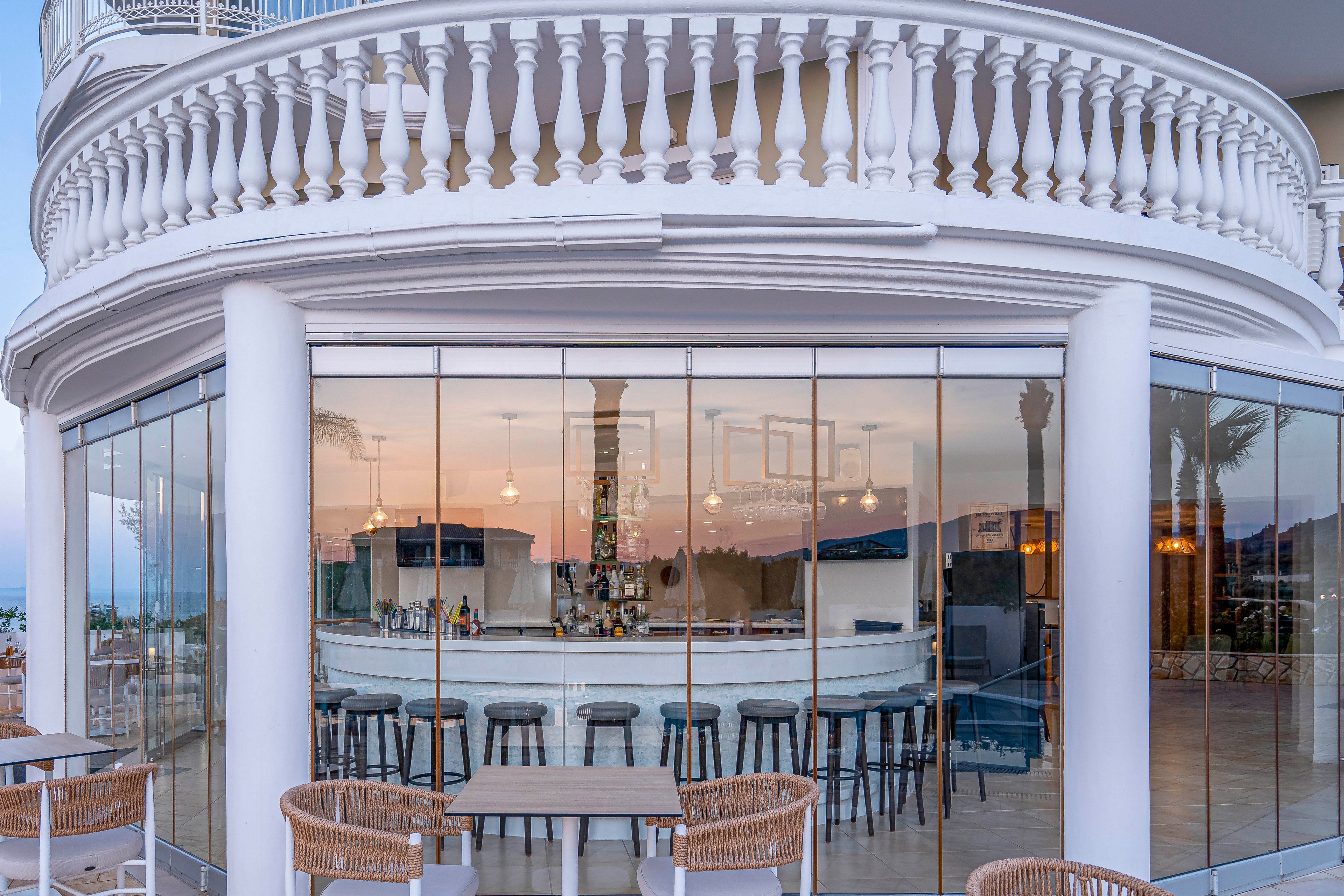 A contemporary bar with a curved glass facade is shown beneath an ornate white balcony and columns. Inside, a sleek counter with bar stools faces a well-stocked back bar, reflected in the large windows. Outdoor tables and wicker chairs are arranged on the terrace, suggesting an inviting place to relax. The soft evening light and pastel sky create a calm, coastal atmosphere.