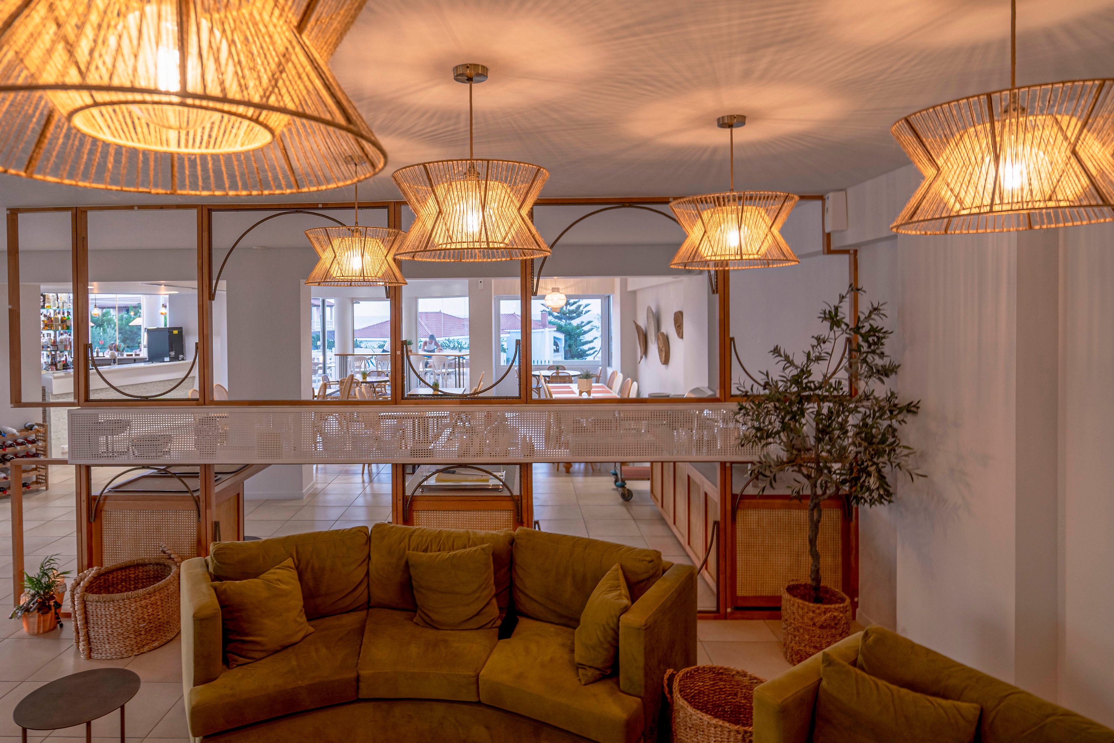 A contemporary lounge area features a curved green sofa arranged around small side tables. Above, several woven pendant lights cast a warm, ambient glow across the room. Large mirrors and windows in the background reflect natural light and reveal a bright dining area beyond. Potted plants and wicker baskets add natural texture and a relaxed, inviting atmosphere.