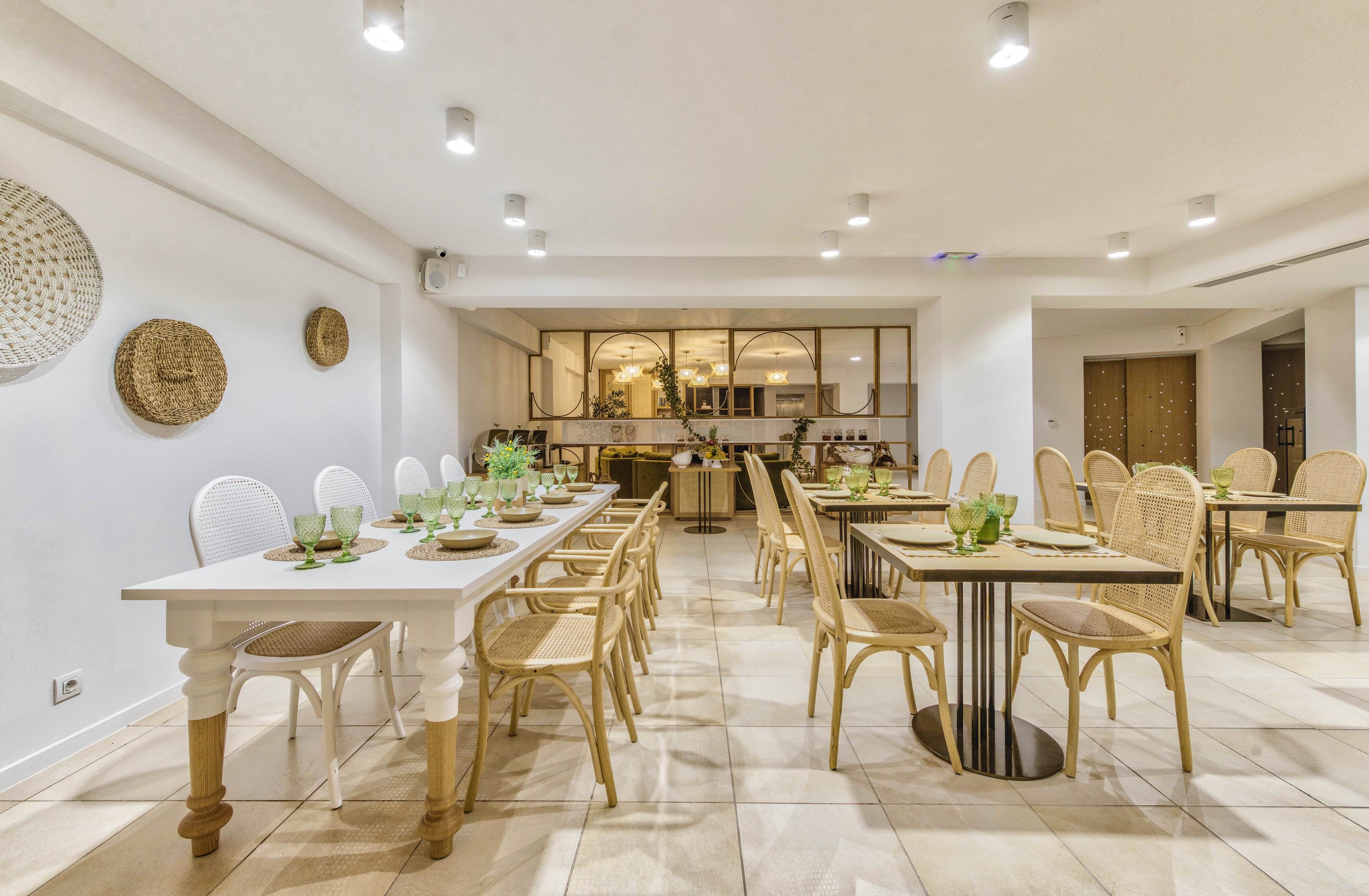 A spacious, modern dining room features long tables set with glassware and greenery. Light wooden wicker chairs line the white and black tables, creating a cohesive and airy look. The room is illuminated by multiple ceiling spotlights and warm ambient lighting from the open kitchen area in the background. Neutral tones, woven wall decor, and tiled flooring contribute to a clean, contemporary atmosphere suitable for gatherings or events.