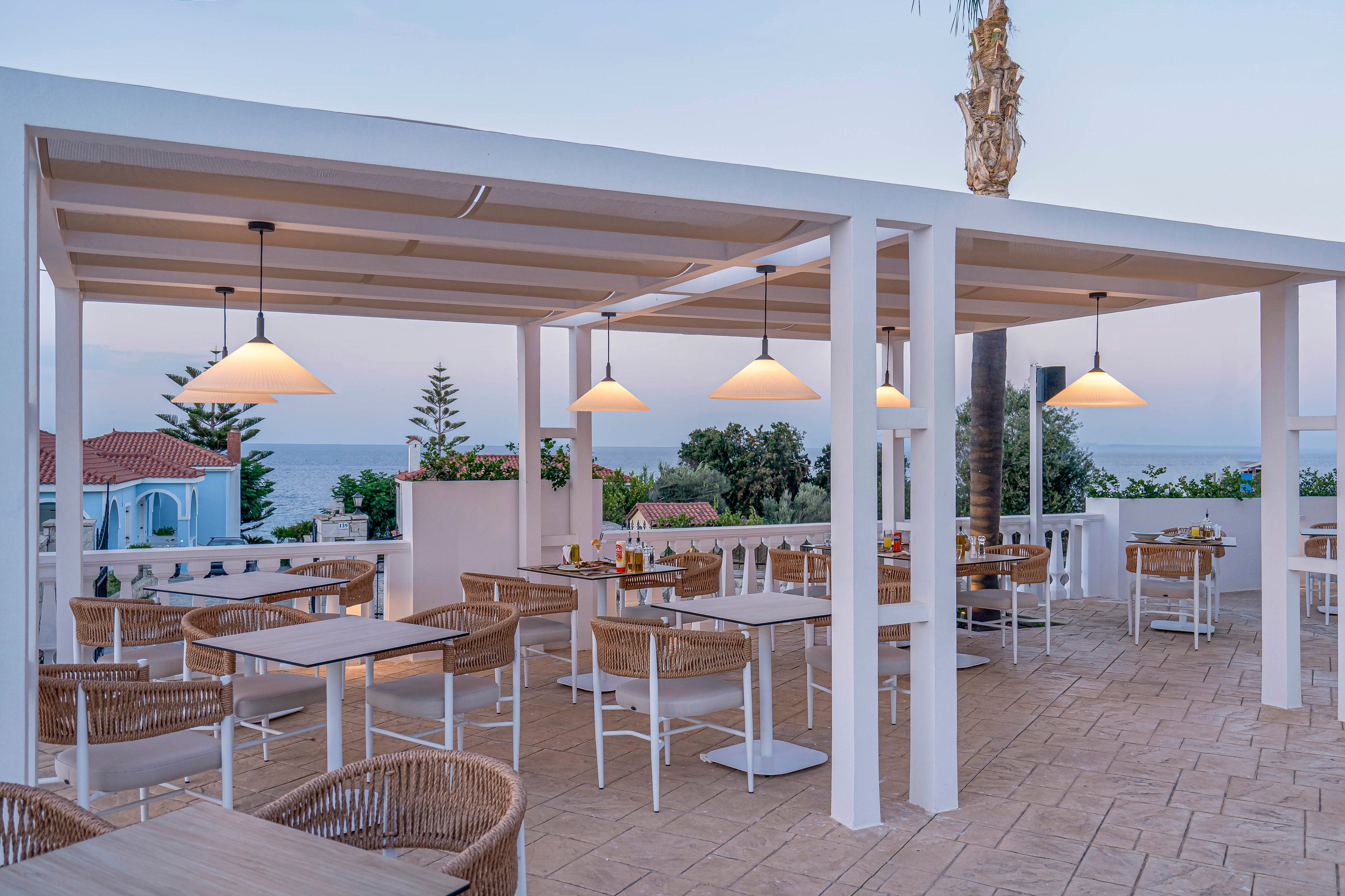 An open-air terrace restaurant features modern white pergolas and wicker chairs overlooking the sea. Several diners sit at tables enjoying a relaxed evening meal as the sun sets. Warm pendant lights hang from the ceiling, casting a soft glow over the tiled floor and minimalist decor. The calm ocean and pastel sky create a tranquil coastal atmosphere.