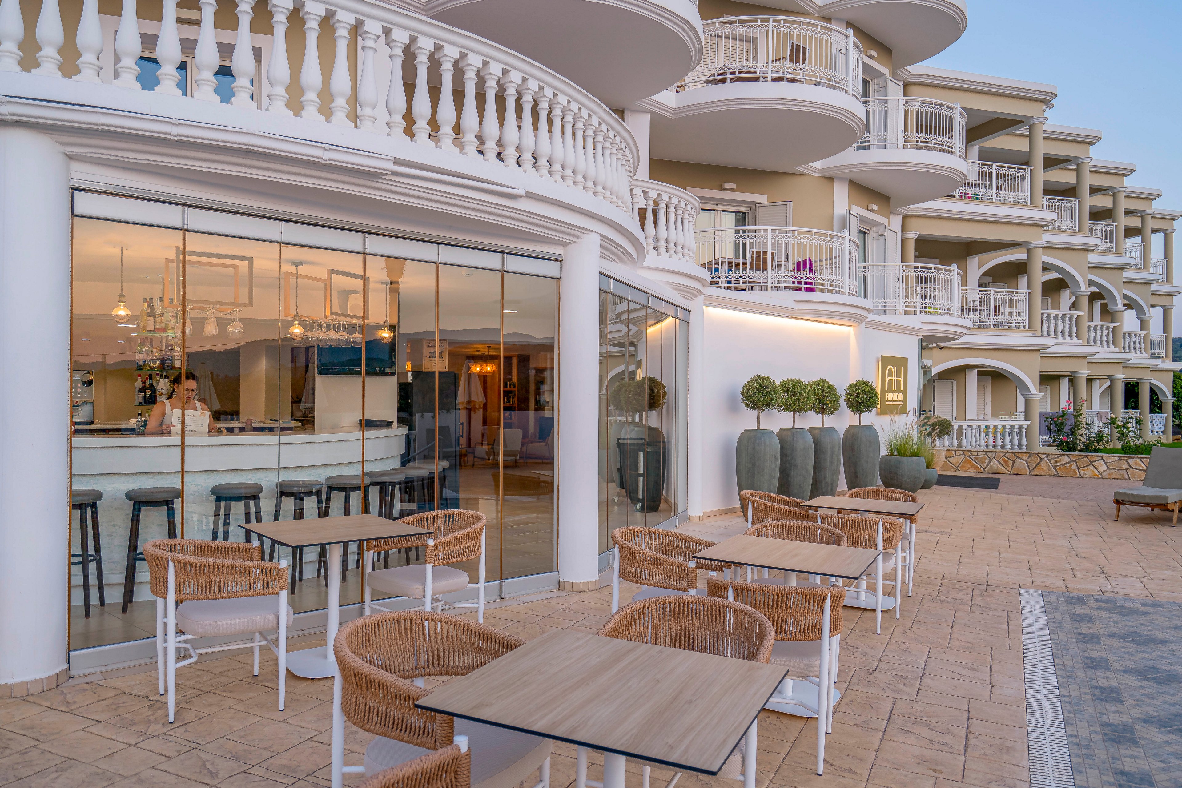 An elegant hotel terrace features outdoor seating with wicker chairs and wooden tables. Behind the glass wall, a bright bar area with high stools and chandeliers is visible. The building showcases curved balconies, white railings, and soft beige tones, suggesting a coastal or resort setting. Neatly trimmed topiary plants and large decorative pots line the walkway, enhancing the upscale atmosphere.