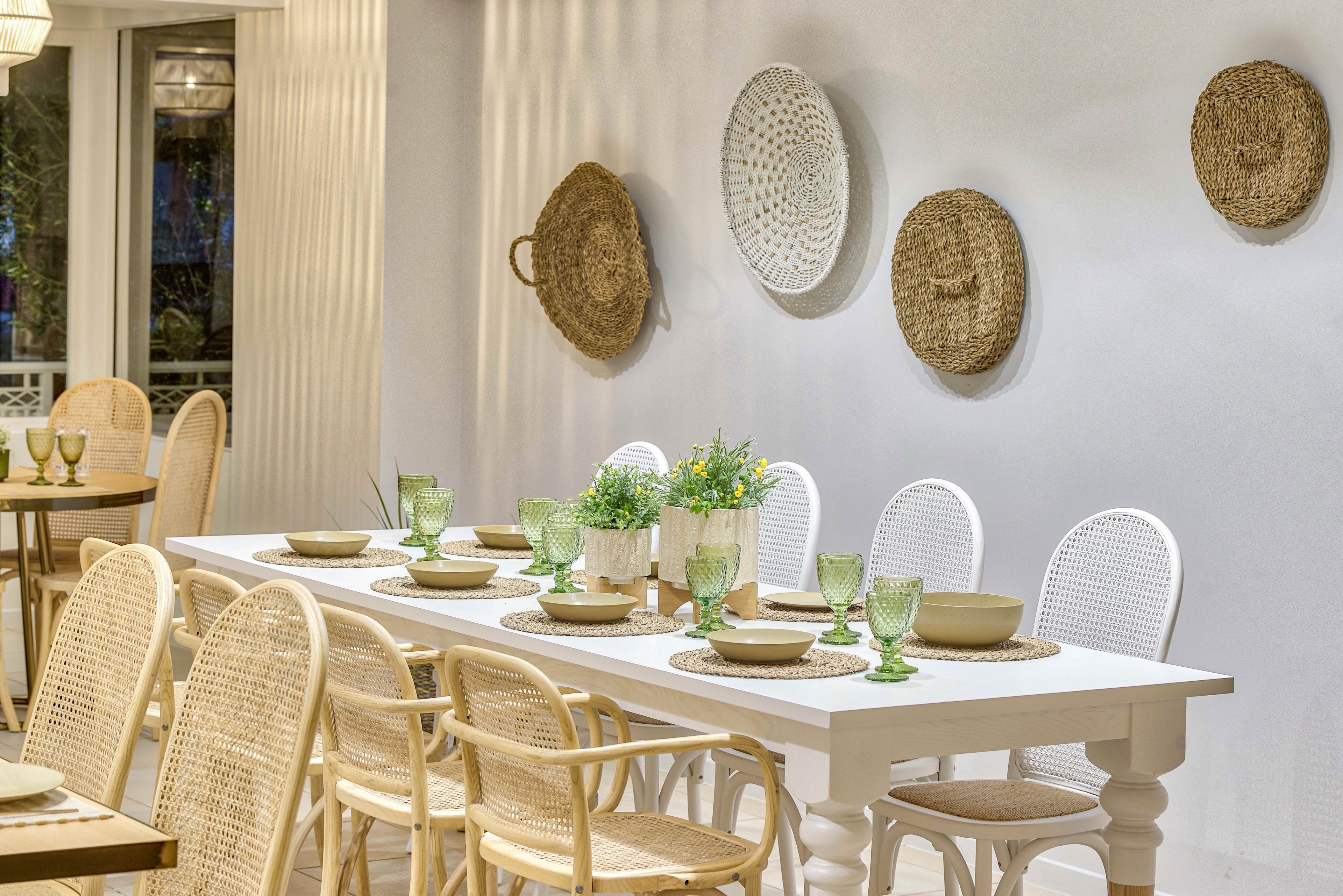 A long white dining table is set with plates, bowls, and green glassware in a bright, cozy room. Wicker chairs and white chairs line both sides of the table, creating an inviting space for gatherings. Decorative woven baskets hang on the light-colored wall, adding texture and a natural touch. Soft lighting and neutral tones give the scene a warm, relaxed atmosphere.