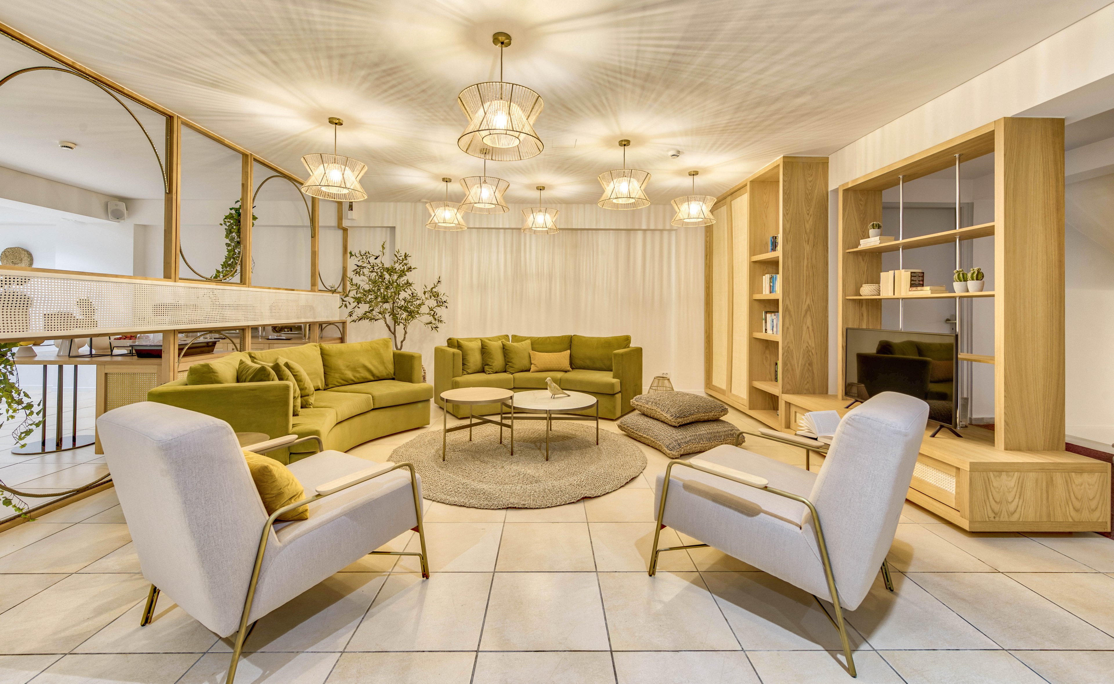 A contemporary lounge features a central seating area with green sofas and light upholstered armchairs arranged around round coffee tables. The room is bright and spacious, with large floor tiles and sheer curtains covering the back wall. Multiple pendant lights hang from the ceiling, casting warm illumination over the space. Built-in wooden shelving and decorative mirrors add a stylish, minimalist touch to the interior.