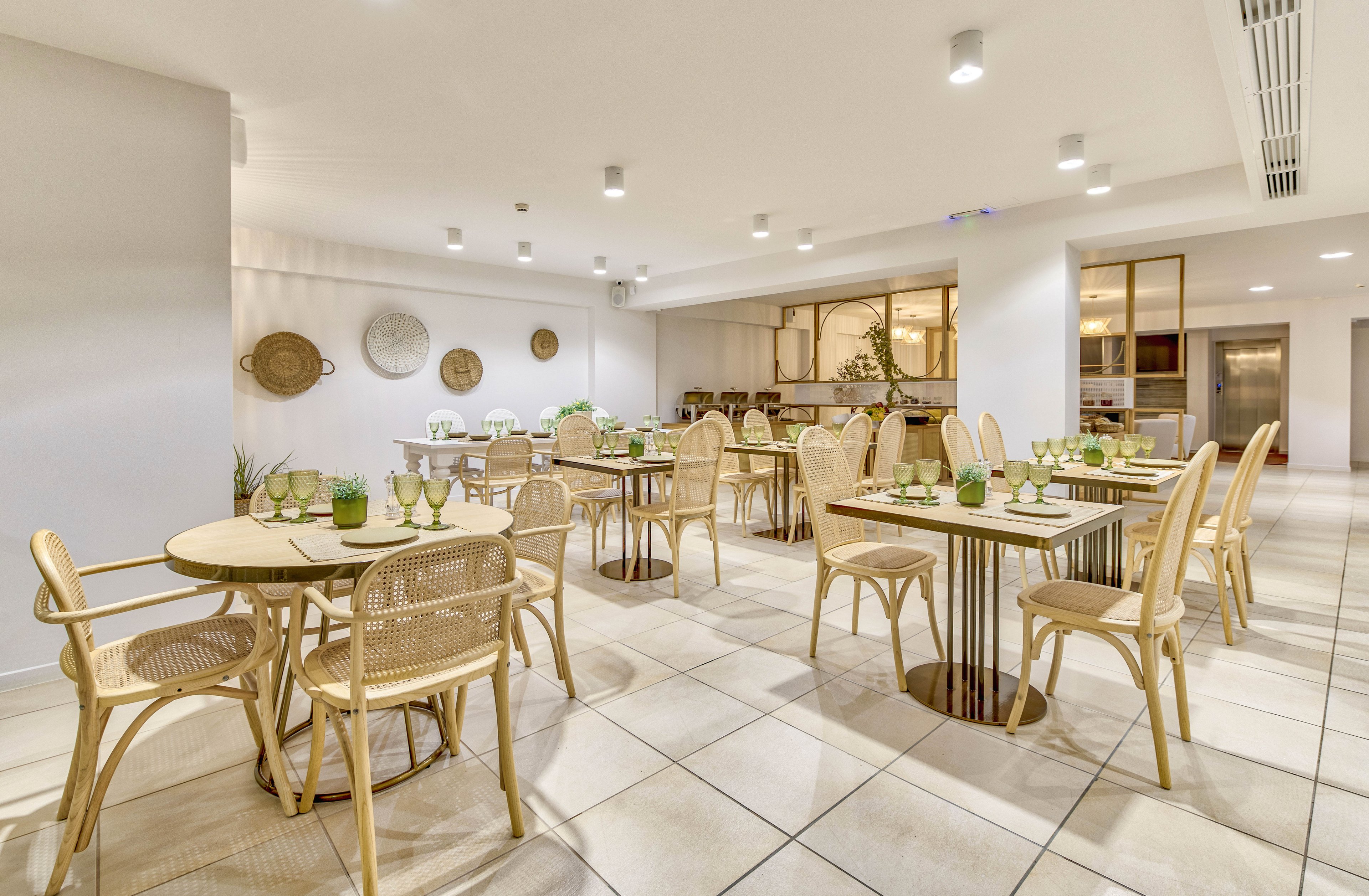 A contemporary restaurant dining area features light wicker chairs and round tables neatly arranged on a tiled floor. The space is brightly lit with recessed ceiling lights and decorated with wall-mounted baskets and minimalist accents. Green glassware and neatly set place settings add a fresh, inviting touch to the neutral color palette. Large windows and open shelving in the background contribute to an airy, modern atmosphere.