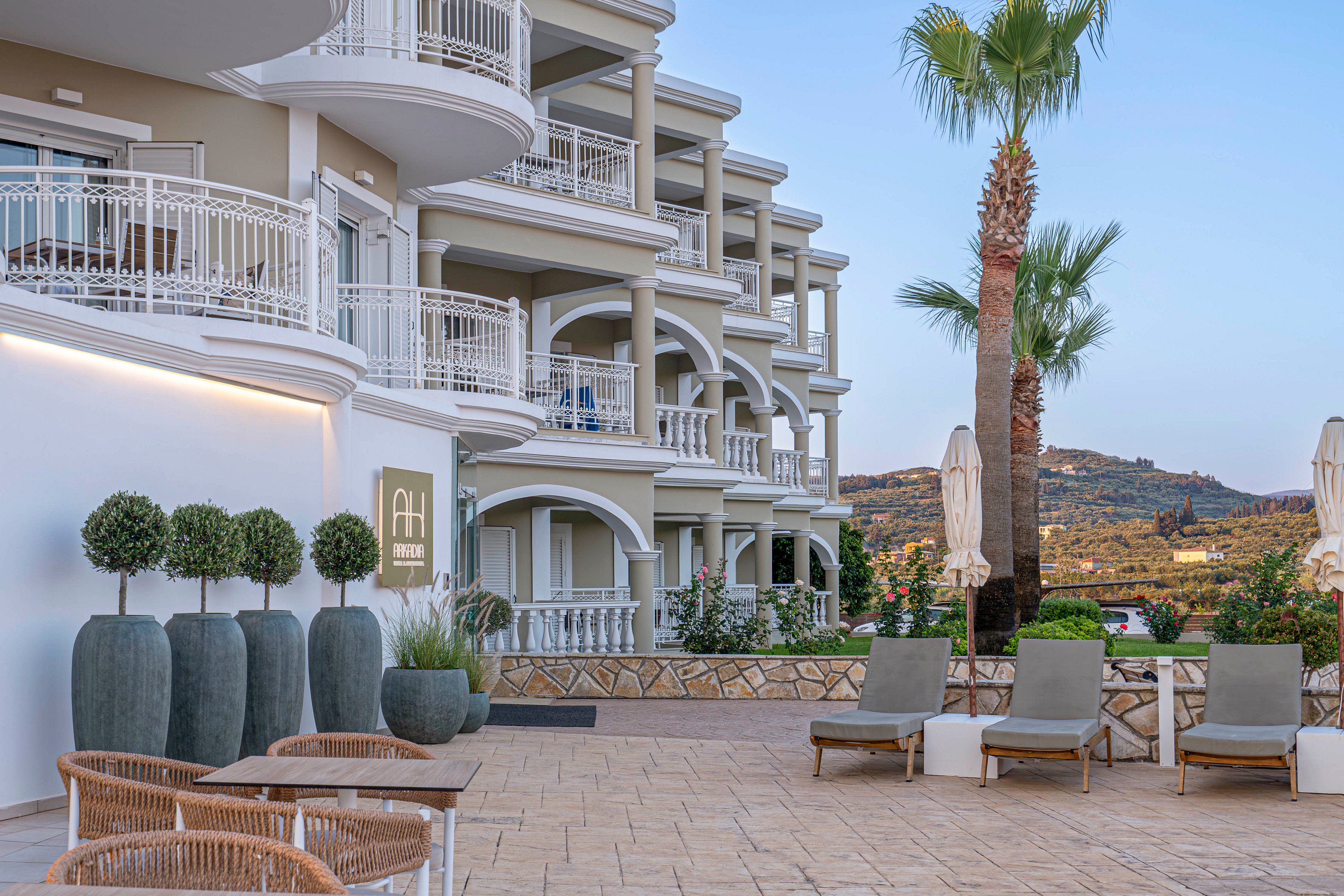A modern multi-story hotel with curved balconies overlooks a spacious outdoor terrace. The foreground features wicker chairs, sun loungers, and large decorative planters with manicured shrubs. A tall palm tree and closed parasols frame the view toward distant hills under a clear evening sky. Soft natural light and neutral tones create a calm, upscale resort atmosphere.