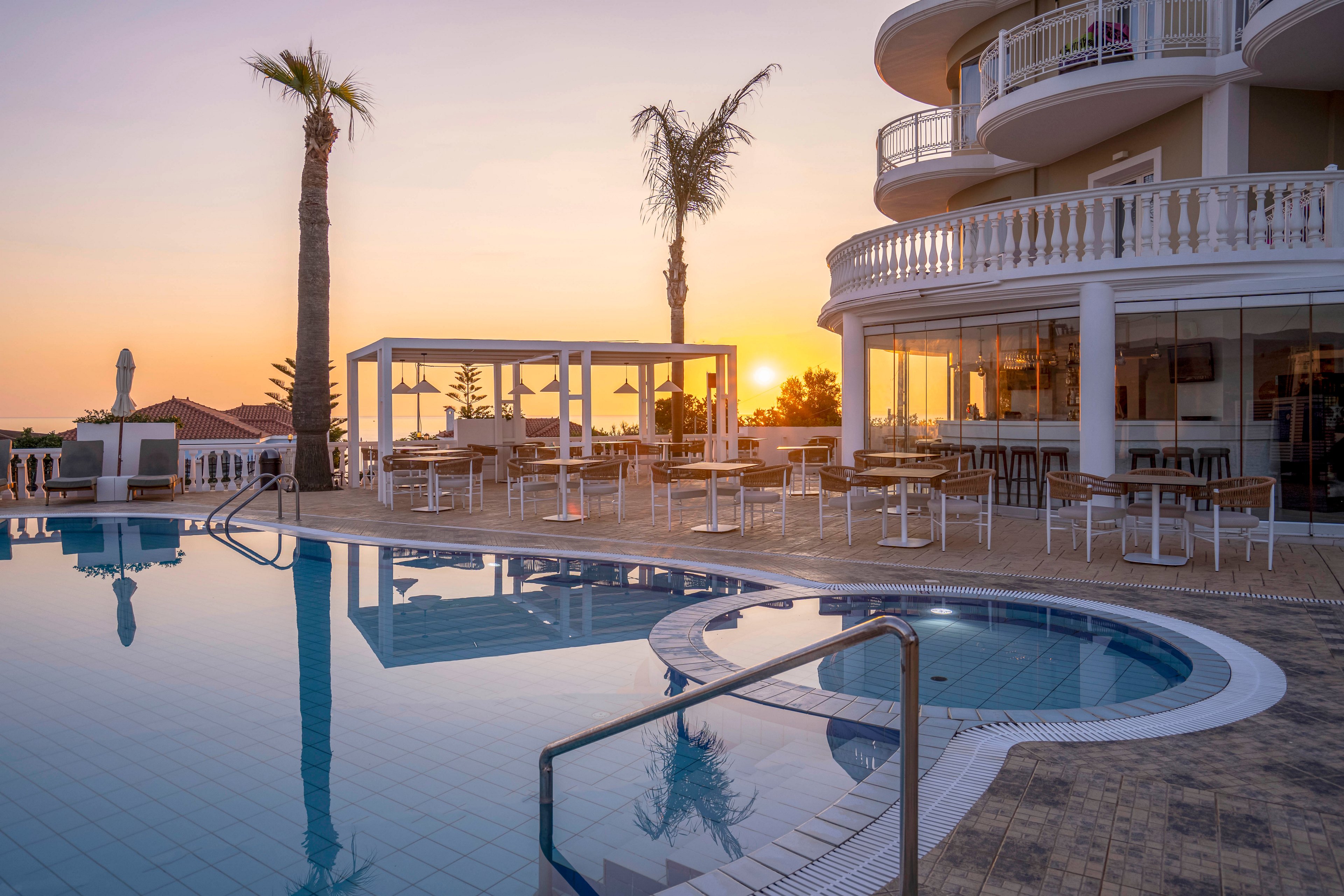 A luxurious hotel pool area is shown at sunset, with calm water reflecting the warm sky. The curved building features balconies and white balustrades overlooking the pool. An open-air terrace with tables, chairs, and pergola structures sits beside the water, ready for guests. Palm trees and distant rooftops frame the horizon as the sun sets, creating a tranquil vacation atmosphere.