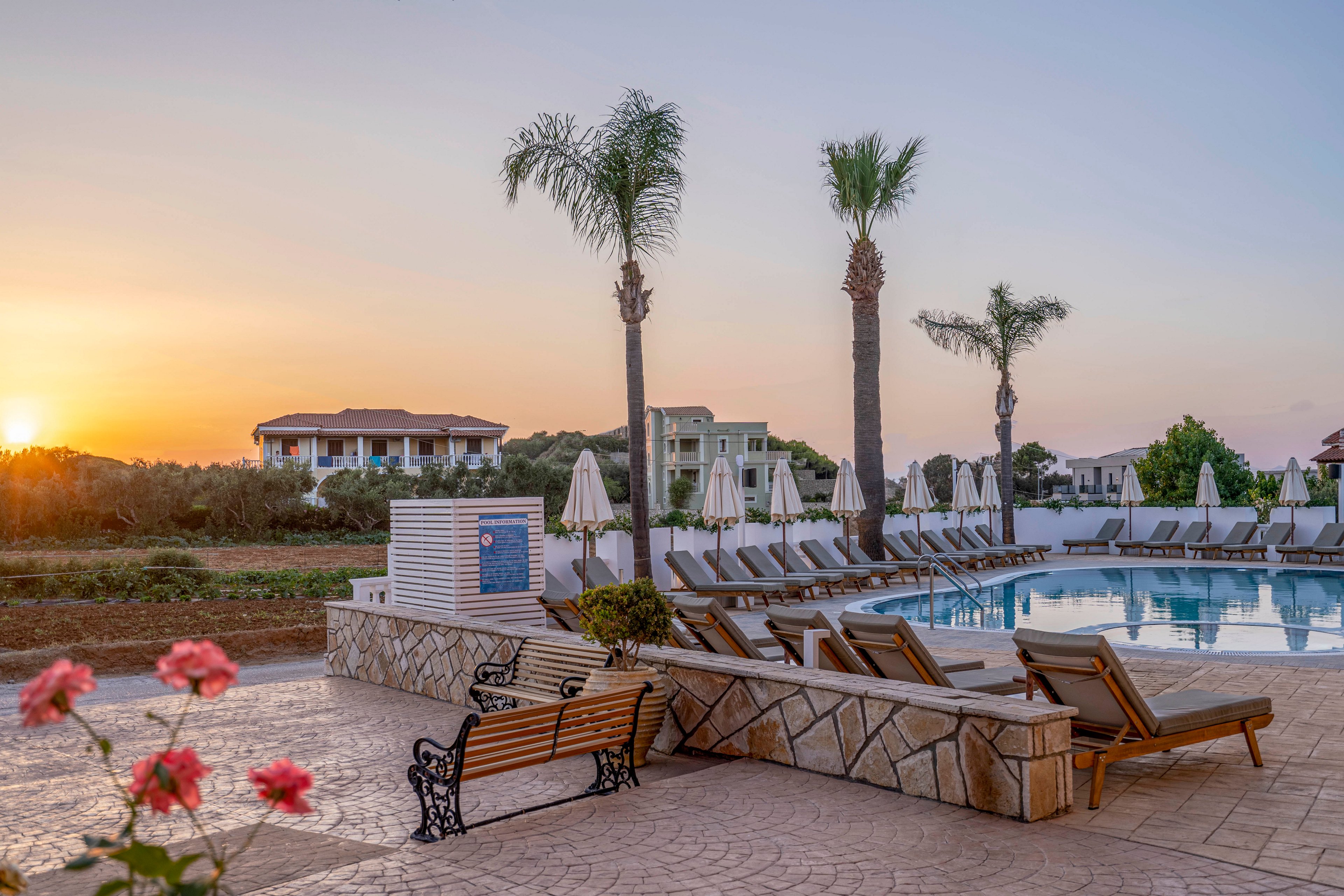 A tranquil resort swimming pool is lined with empty sun loungers and umbrellas under tall palm trees at sunset. The scene overlooks landscaped grounds and low-rise hotel buildings in the distance. Warm evening light reflects off the calm water, creating a peaceful vacation atmosphere. A stone patio with benches and blooming flowers frames the foreground, enhancing the relaxed, tropical feel.