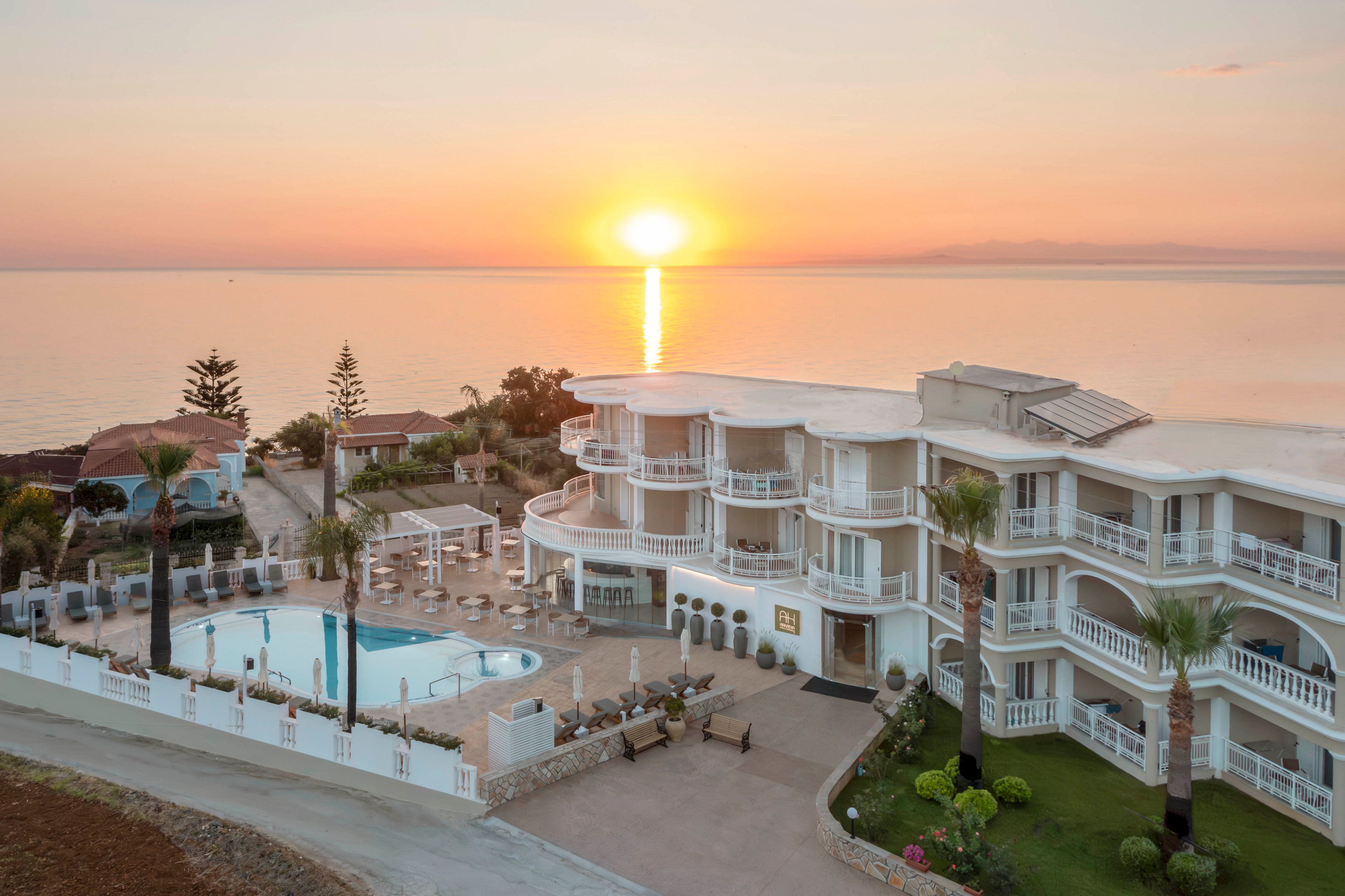 A coastal resort hotel is captured from an elevated viewpoint as the sun rises over a calm sea. The multi-story white building features curved balconies and Mediterranean-style architecture. A swimming pool and sun loungers sit within a fenced terrace surrounded by palm trees and landscaped greenery. Warm pastel colors from the sunrise reflect on the water, creating a tranquil vacation atmosphere.