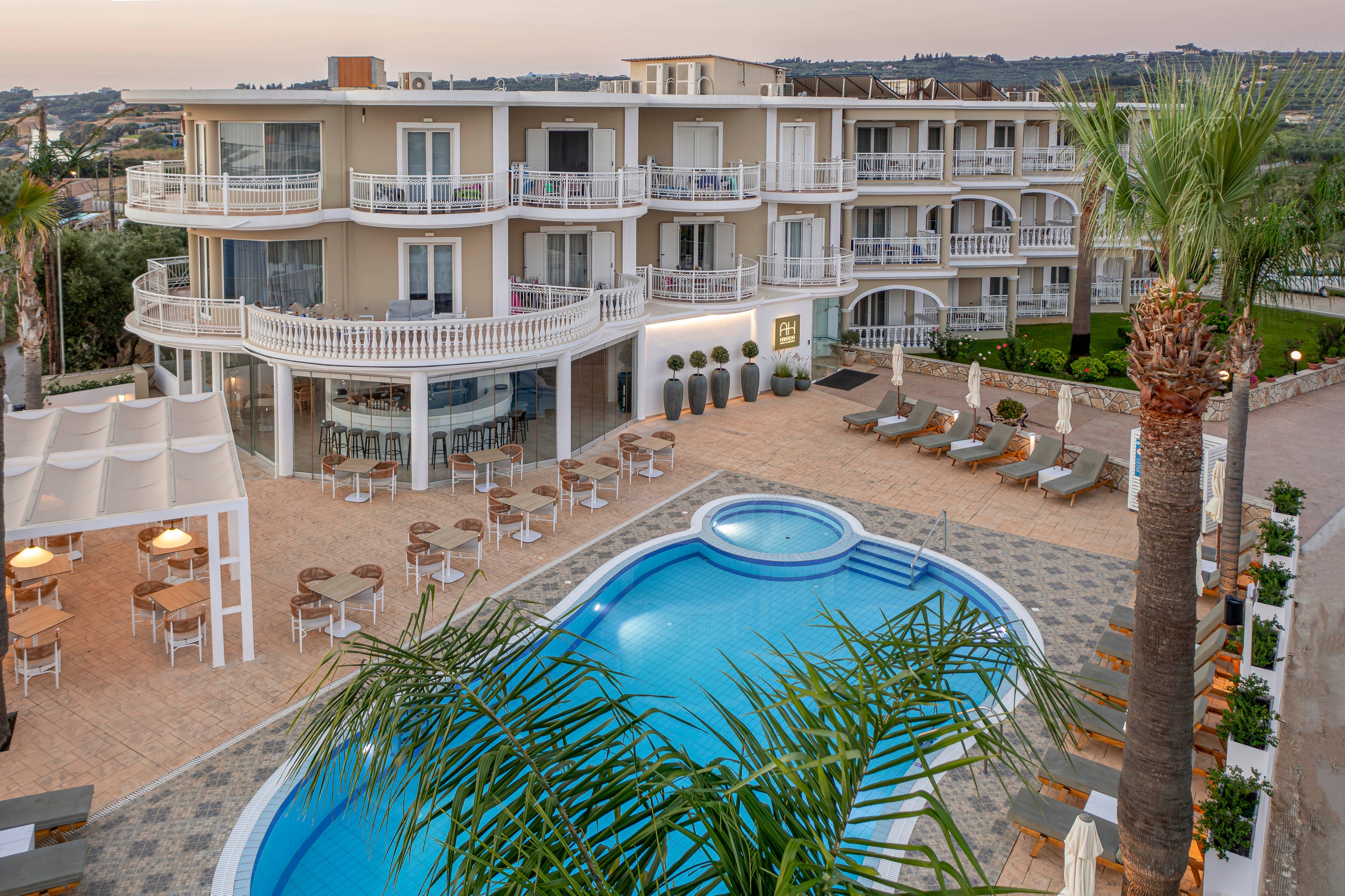 A multi-story resort hotel with curved balconies overlooks a central swimming pool area. The scene is set at sunset, casting warm light over the building and surrounding landscape. Sun loungers and umbrellas line the poolside terrace, while an outdoor dining area with tables and chairs sits under a pergola. Palm trees and distant hills enhance the relaxed, vacation atmosphere of the property.