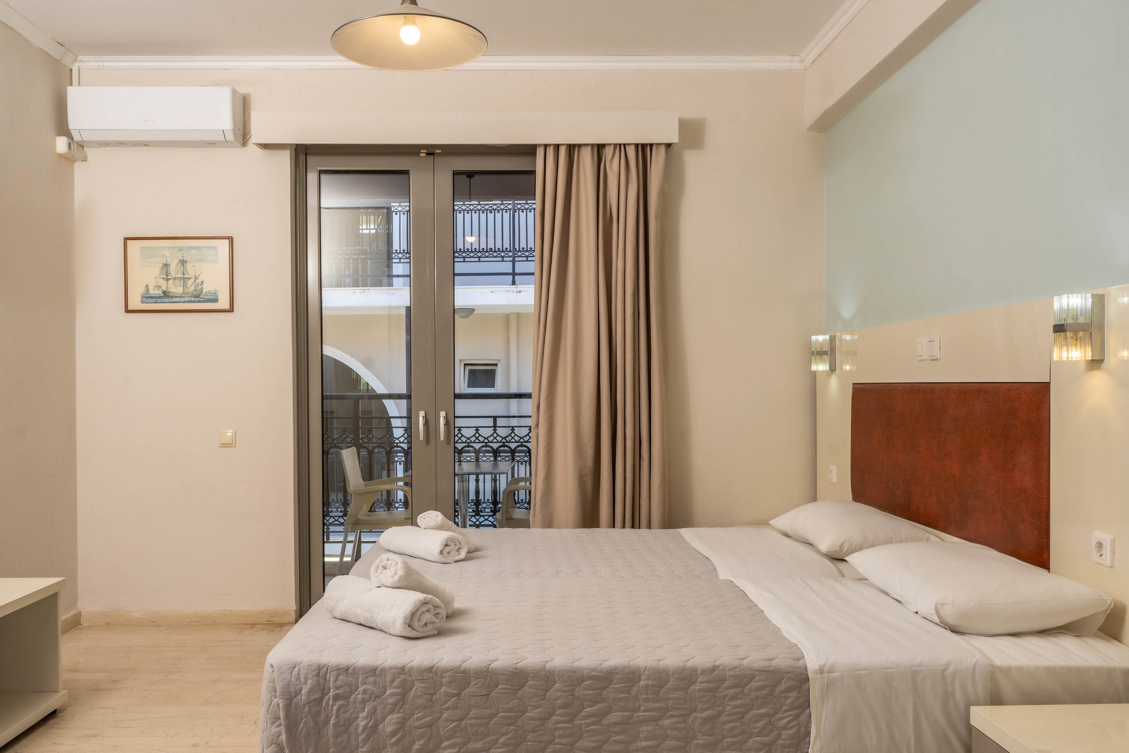 A neatly made double bed with folded towels is the focal point of this modern hotel room. The space features neutral walls, a wooden headboard, and soft lighting from wall sconces and a ceiling lamp. Large glass doors open to a balcony with a view of an adjacent building and railings. Minimal decor and clean lines create a calm, contemporary atmosphere suitable for travelers.