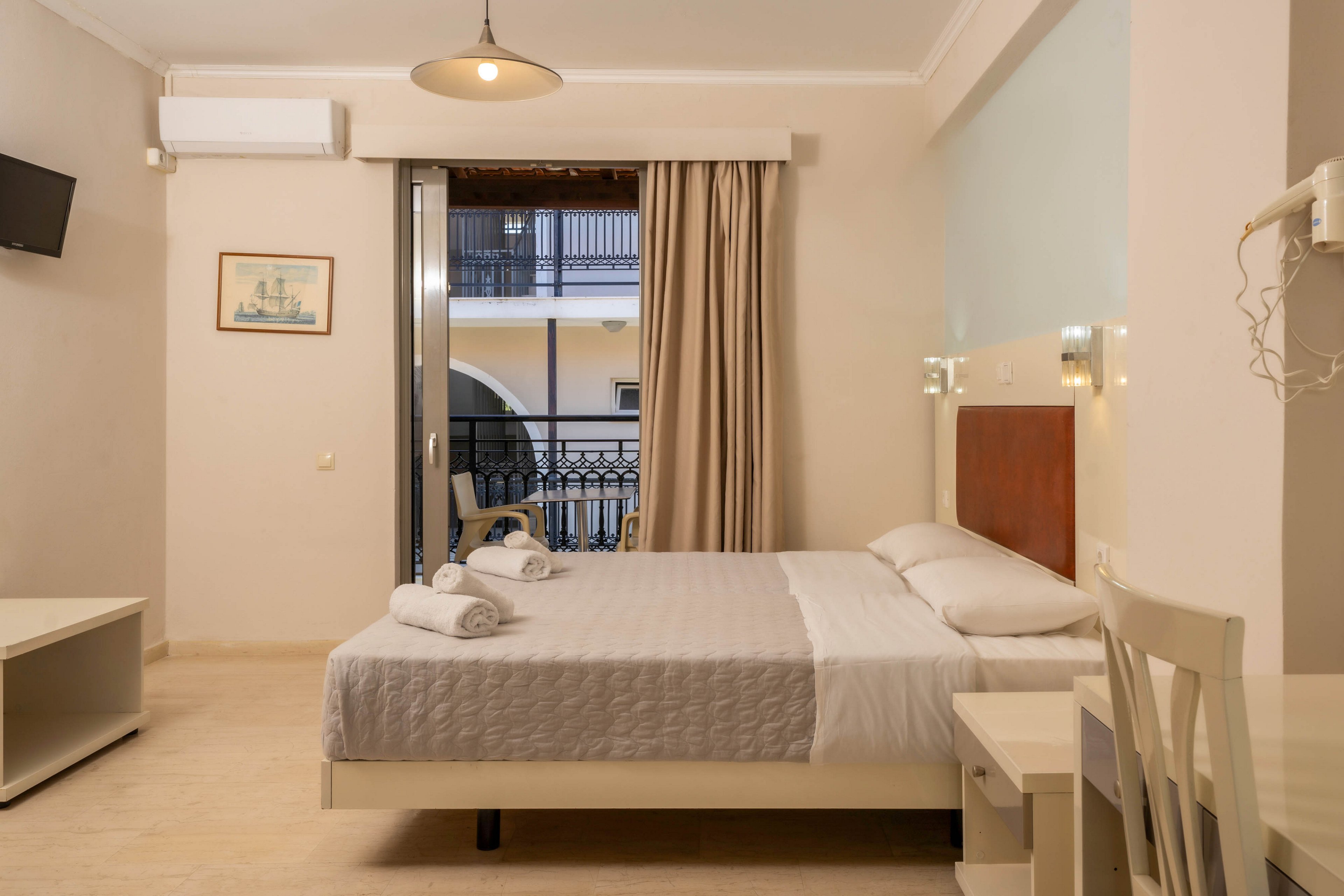 A neatly made double bed with folded towels sits in a modern hotel room. Neutral-colored walls and light wood furniture create a calm, minimalist atmosphere. A sliding glass door opens to a balcony with a view of neighboring building balconies. Soft lighting, wall-mounted lamps, and a small wall-mounted TV complete the simple, comfortable interior.