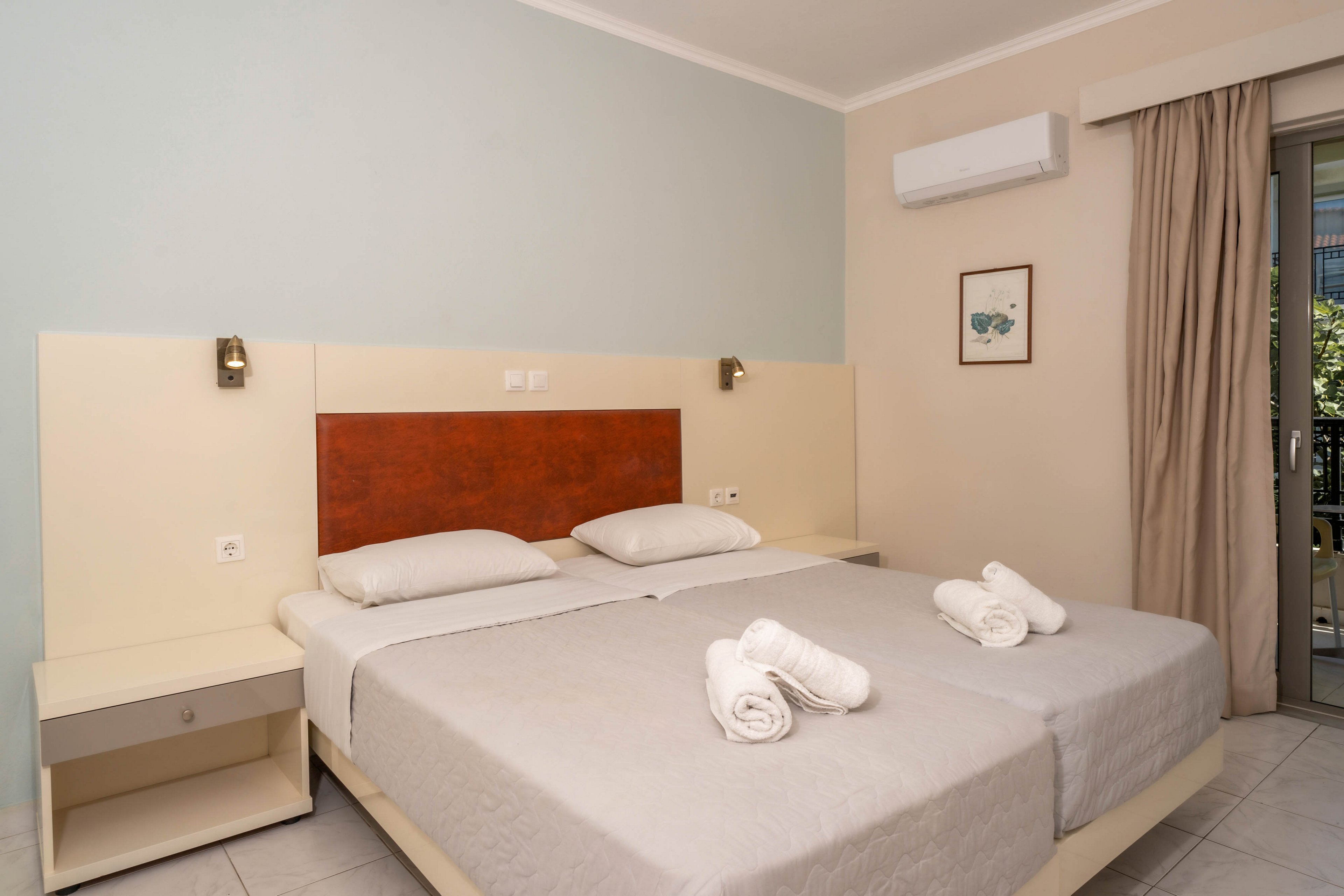 A clean, contemporary hotel room features two twin beds with light gray bedding and rolled towels placed on top. The room is decorated in neutral tones, with a wooden headboard, wall-mounted reading lights, and a framed artwork. An air conditioning unit is mounted above the bed, and a sliding glass door leads to a balcony with outdoor views.