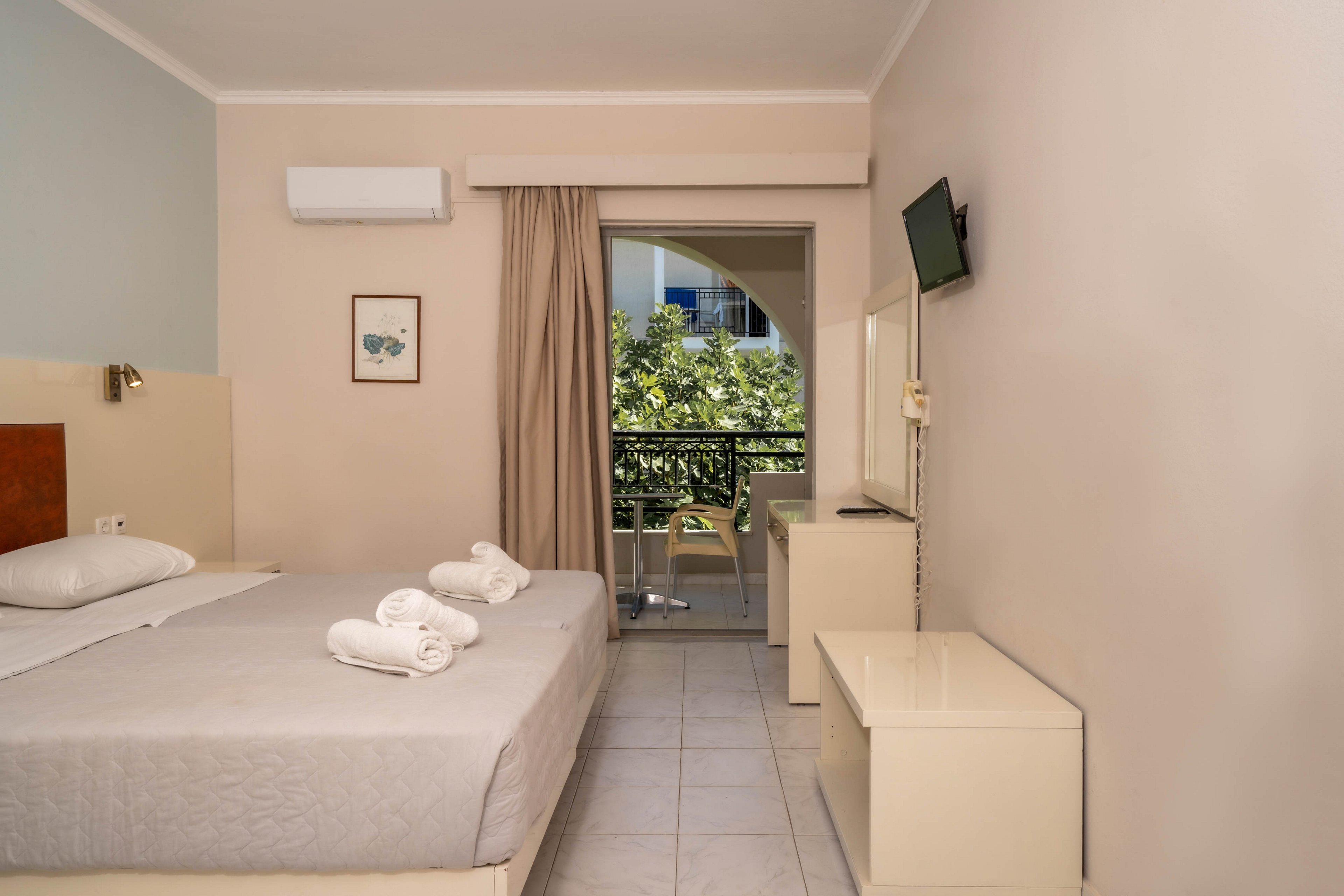 A neatly arranged hotel room features a double bed with folded towels, a wall-mounted television, and a small desk. The space opens to a balcony with outdoor seating, overlooking greenery. The room is decorated in neutral tones with minimal artwork and has a clean, contemporary style. An air conditioning unit is visible above the bed, and natural light brightens the interior.