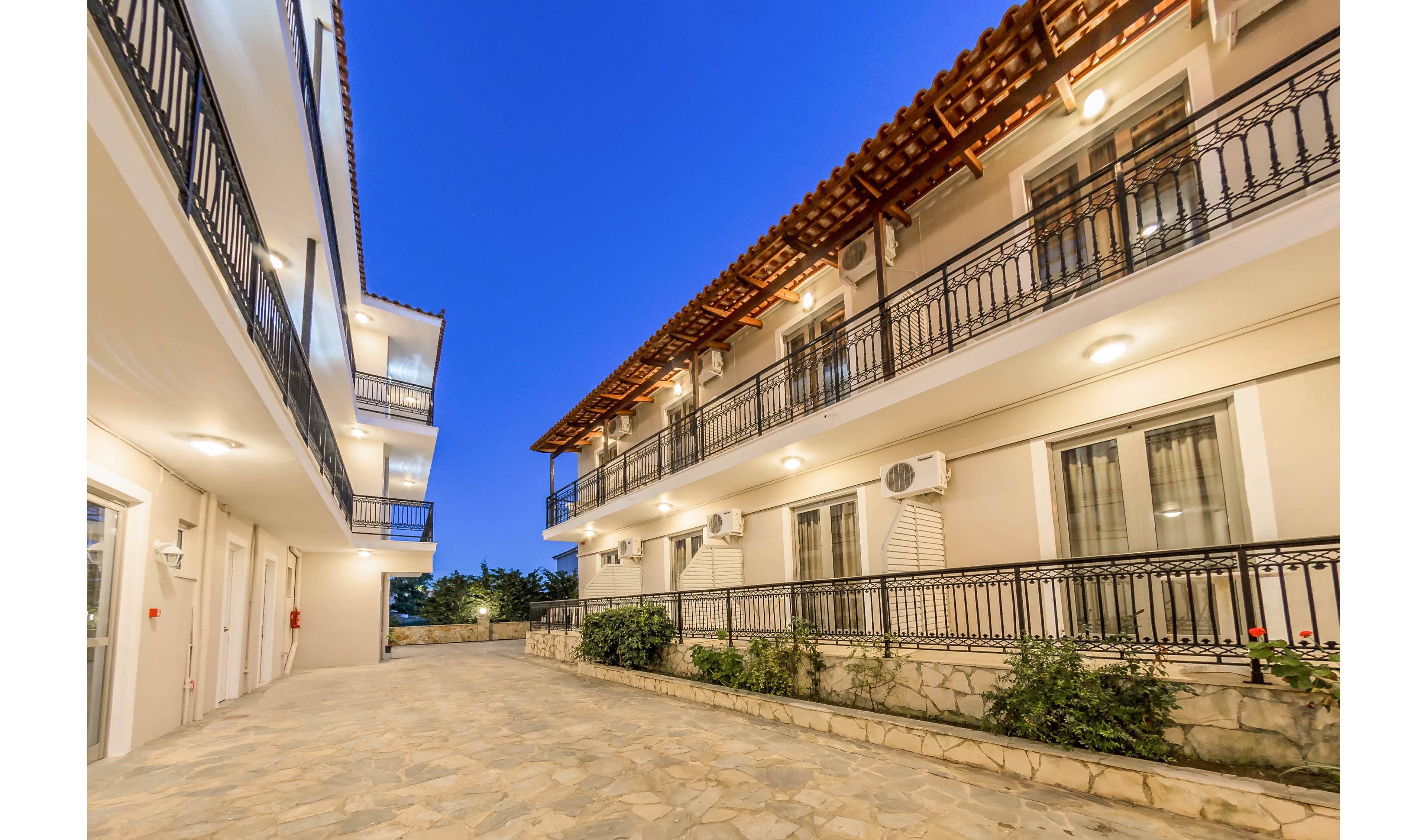 A well-lit courtyard is surrounded by two-story apartment buildings with balconies and exterior lighting. The setting is outdoors during twilight, with a clear blue sky overhead. The architecture features white walls, black railings, and visible air conditioning units. No people or visible text are present in the image.