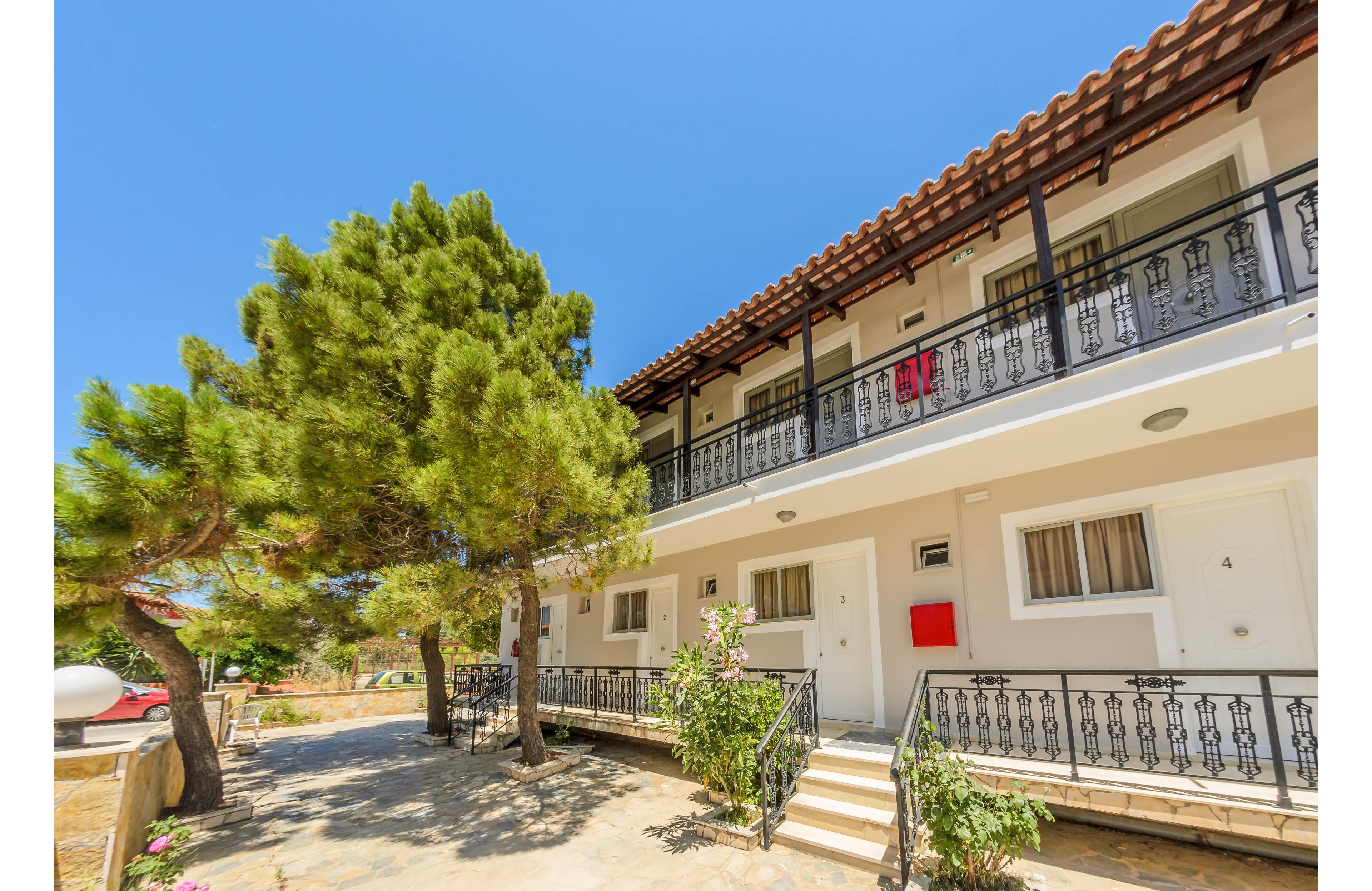 A two-story apartment building features white exterior walls, black railings, and a tiled roof. The setting is outdoors on a bright, sunny day with a clear blue sky, and a large pine tree provides shade near the entrance. Several doors and windows are visible, with a red mailbox and the number 4 clearly marked on one door. The overall mood is relaxed and inviting, with Mediterranean architectural influences.