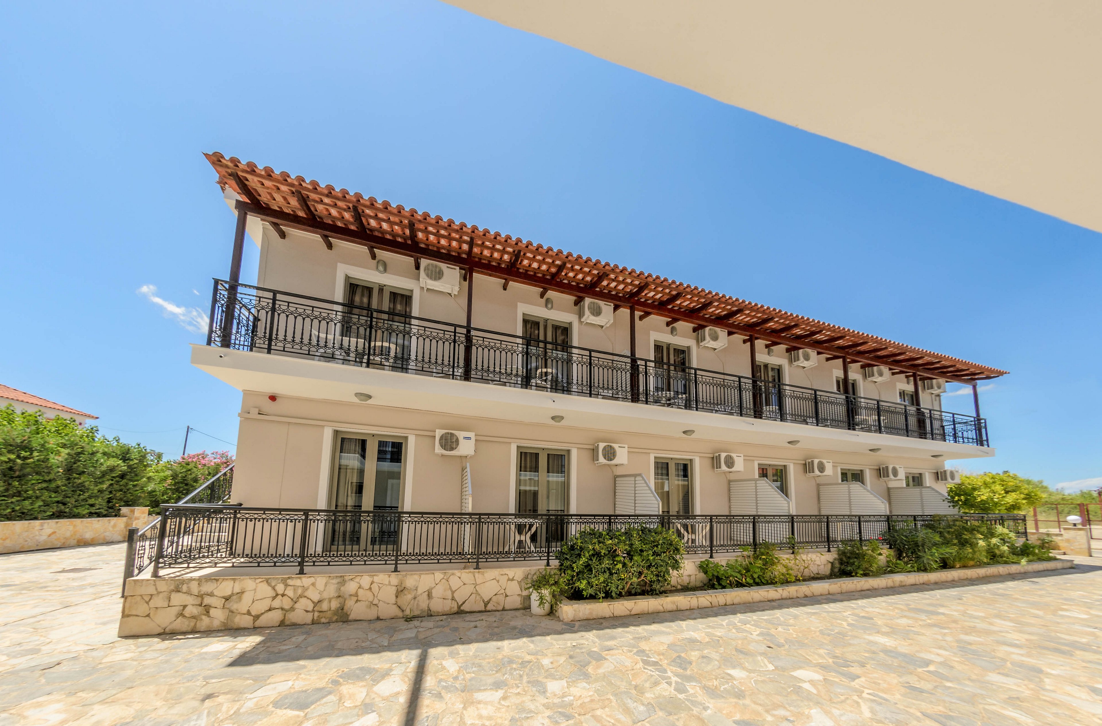 A contemporary two-story apartment building features multiple balconies and air conditioning units on each unit. The structure is set in a sunlit, open courtyard with stone paving and minimal landscaping. The building's exterior is painted in neutral tones, complemented by a red-tiled roof and black railings. The scene is captured under a bright blue sky, evoking a warm and inviting atmosphere. No visible text or numbers are present in the image.