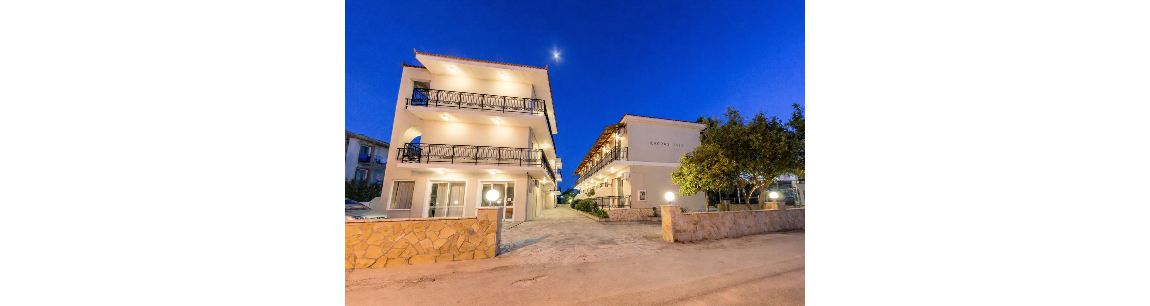 Two modern hotel buildings are brightly illuminated under a deep blue evening sky, with the moon visible above. The setting features clean architectural lines, balconies, and a stone wall at the entrance. The sign 'Karras Livin' is clearly visible on one building, and the scene is tranquil with no people present. Warm lighting and a clear sky create a welcoming atmosphere.