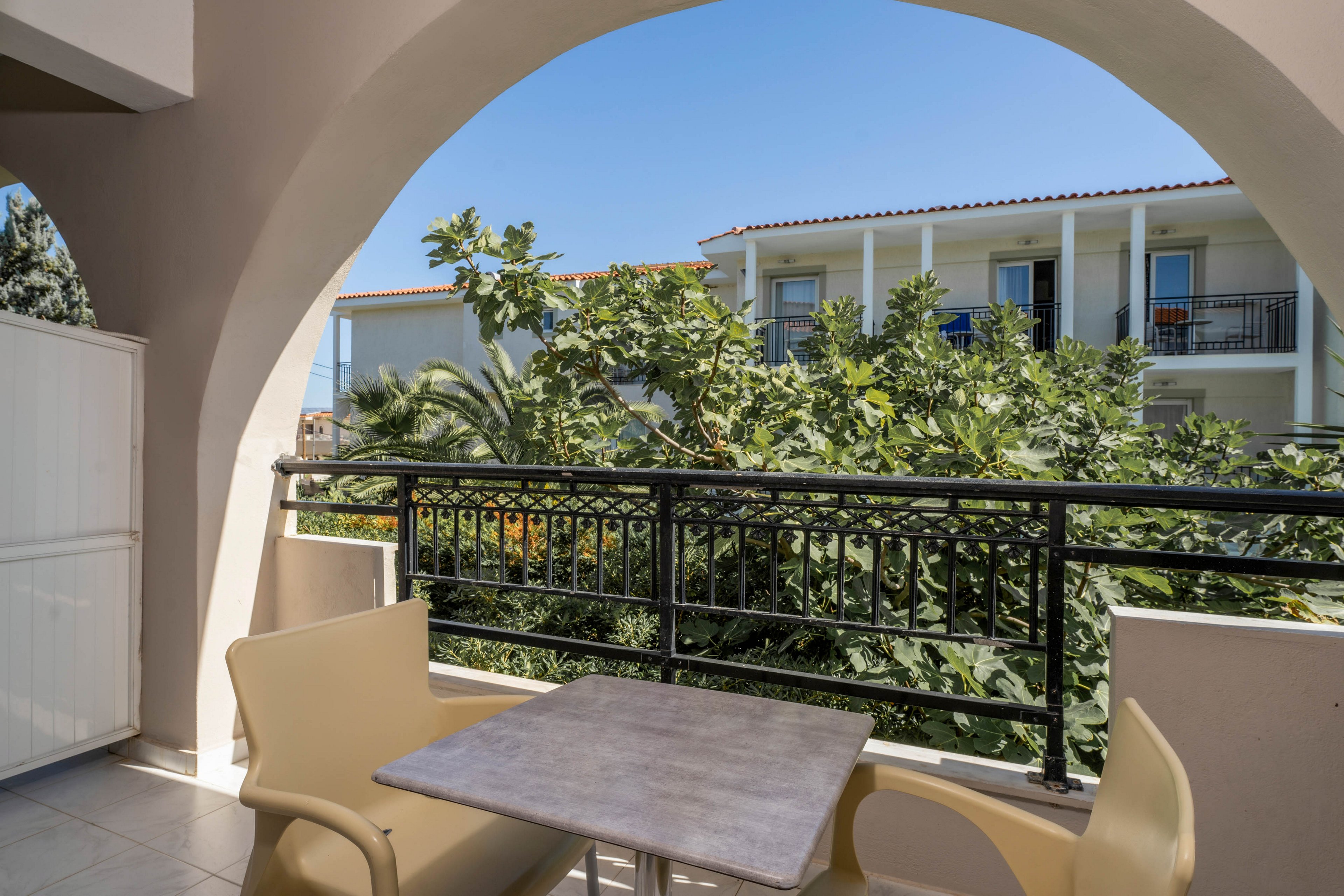 A small outdoor balcony features a square table and two beige chairs, set under an arched opening. The view overlooks lush green foliage and a modern white building with balconies. The scene is brightly lit by natural sunlight, creating a relaxed and inviting atmosphere. The sky is clear and blue, enhancing the summery mood.