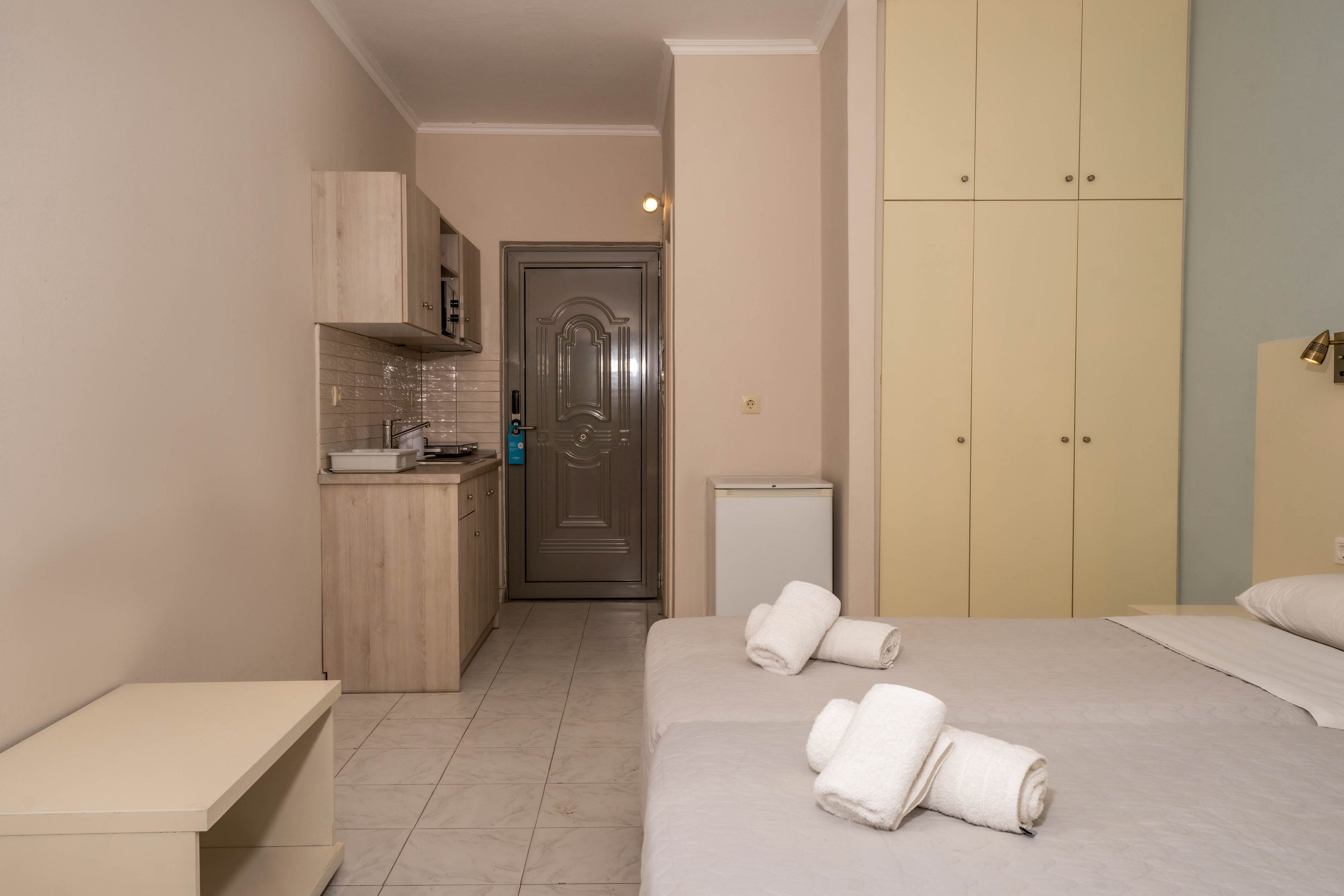 A compact studio apartment features two neatly made twin beds with rolled towels placed on top. The room includes a small kitchenette with light wood cabinets, a microwave, and a sink, set against a tiled backsplash. Neutral tones dominate the space, with cream-colored walls and cabinetry, creating a clean and modern atmosphere. The entry door and ample built-in storage are visible, emphasizing functionality and simplicity.