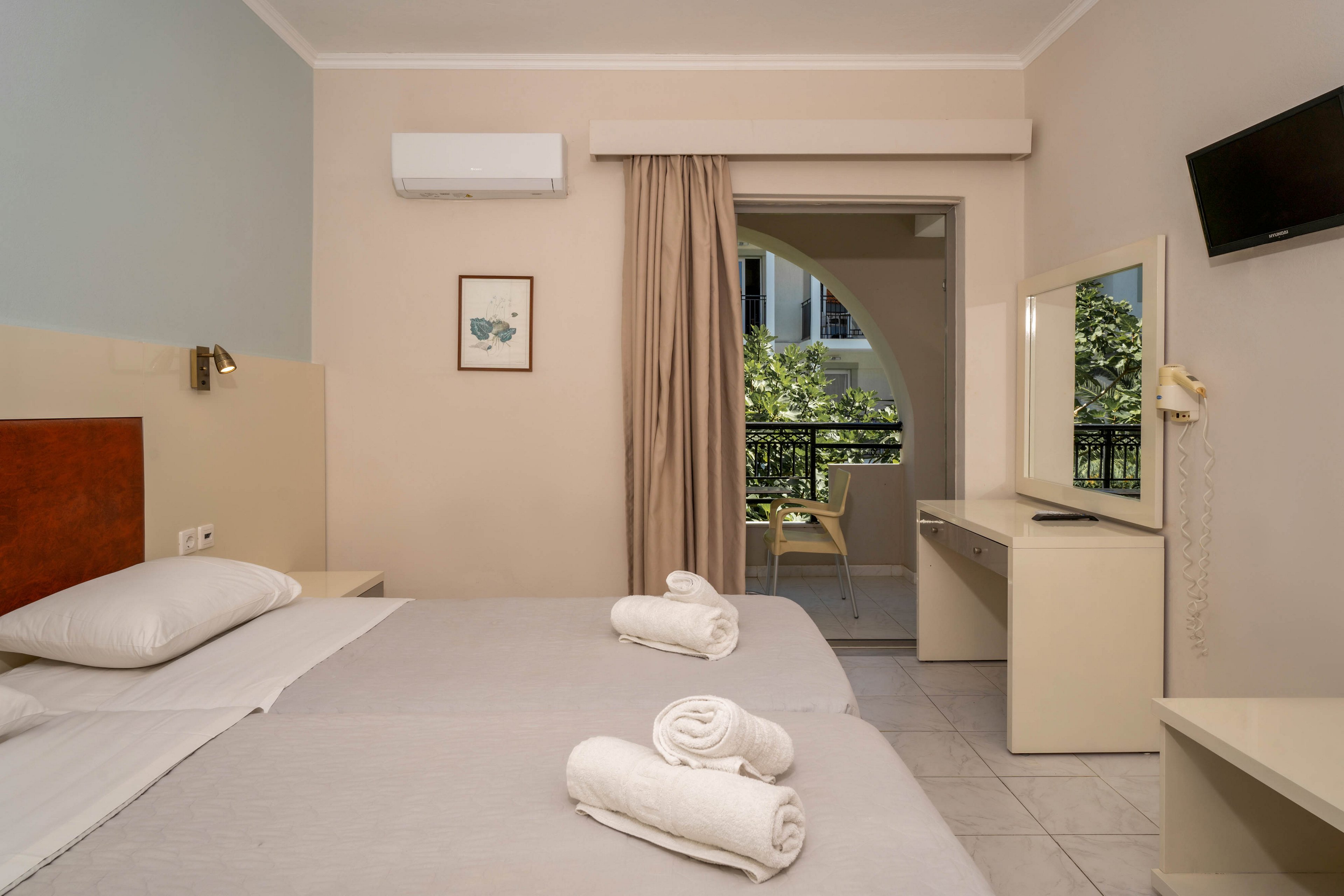A neatly arranged hotel room features a double bed with fresh white towels placed on top. The room includes a wall-mounted television, air conditioning unit, and a desk with a mirror. Natural light enters through a sliding door leading to a balcony with outdoor seating and green foliage visible. The overall palette is neutral with soft beige and white tones.