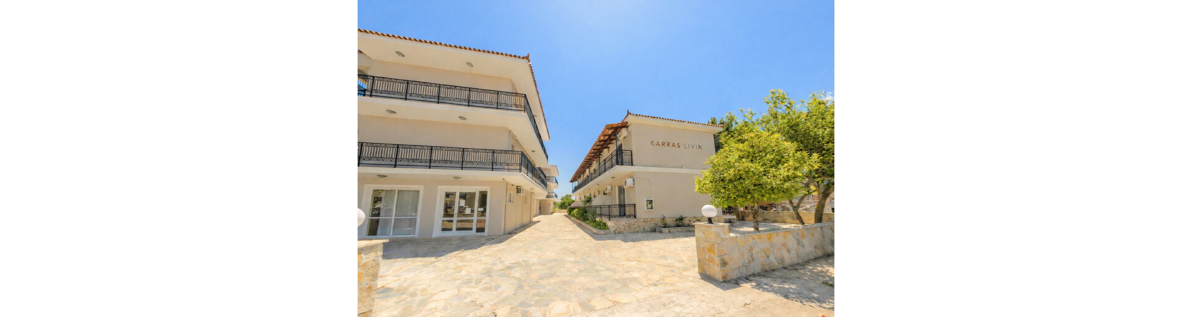 Two modern hotel buildings are shown under a clear blue sky, with the name 'Karras Livin' visible on one structure. The scene features stone walkways, balconies, and landscaped greenery, creating a welcoming and relaxed atmosphere. The bright sunlight and open courtyard enhance the Mediterranean architectural style.
