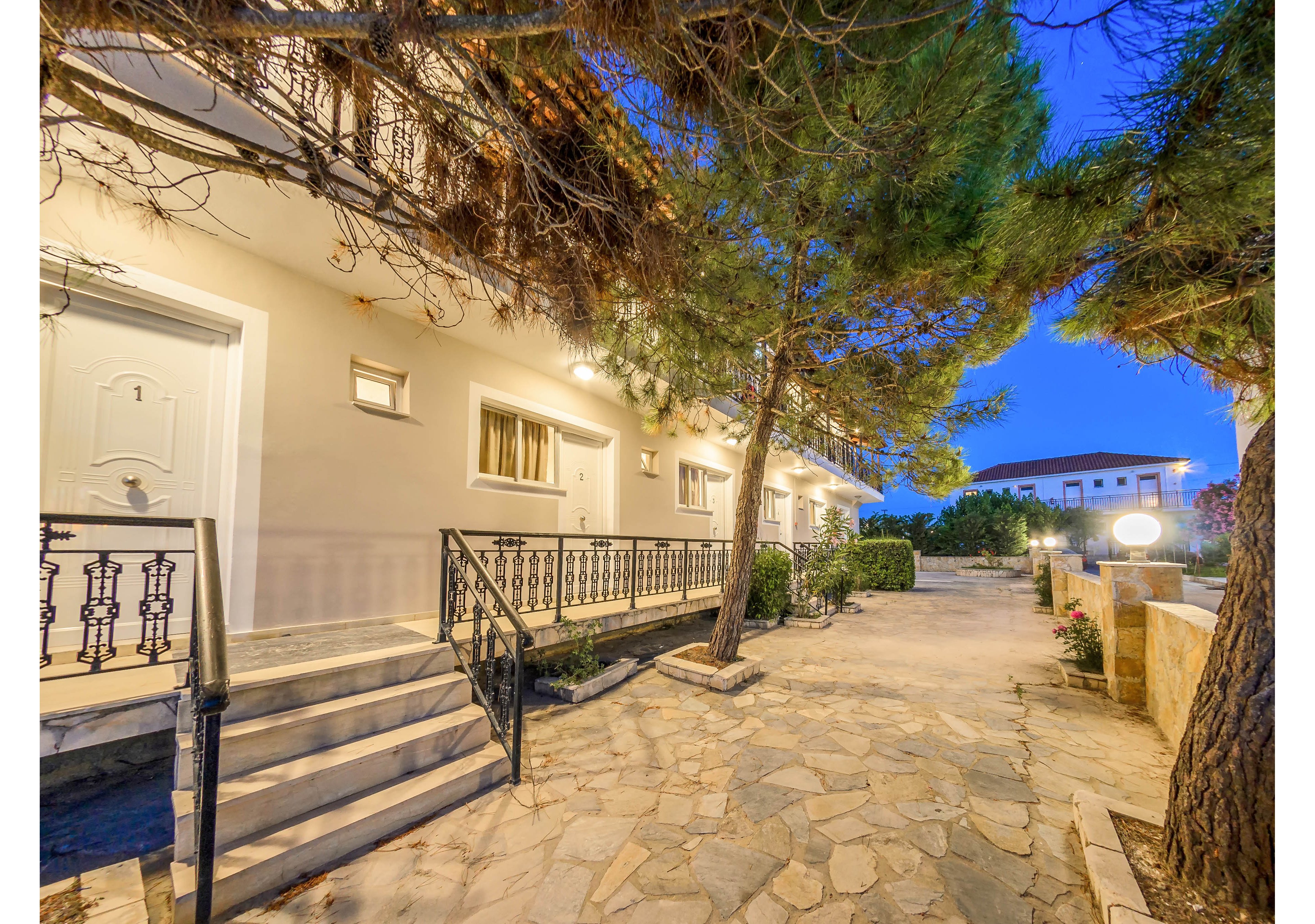 A well-lit exterior of an apartment building is shown at dusk, featuring a white door marked with the number '1'. The setting includes stone steps, a paved walkway, and lush trees providing shade. Warm lighting and a tranquil atmosphere highlight the residential environment. The scene is empty of people, emphasizing the peaceful outdoor space.