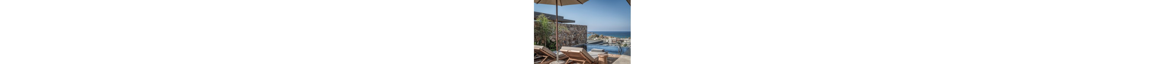 A modern seaside resort features wooden sun loungers beside an infinity pool overlooking the ocean. Stone walls and contemporary architecture frame the tranquil outdoor terrace. An umbrella provides shade while folded towels rest on the loungers, suggesting relaxation and leisure. In the distance, coastal buildings and rocky shoreline meet the calm blue sea under a clear sky.