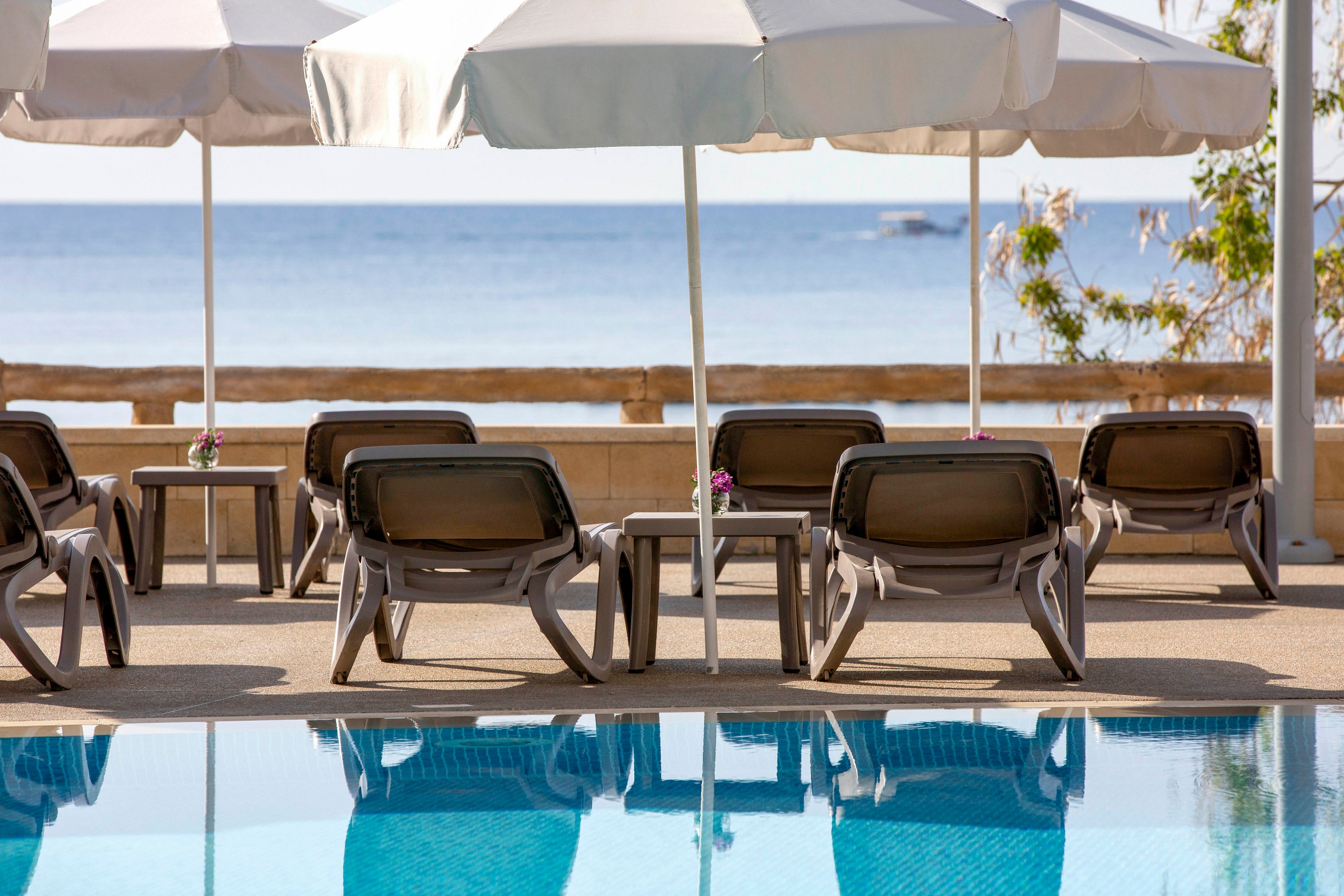 A row of empty sun loungers sits beside a clear blue swimming pool, facing toward a calm sea. Large white umbrellas provide shade over the chairs, suggesting a relaxing resort setting. The background features a wooden railing and distant water horizon under a bright sky. The overall scene conveys tranquility, leisure, and vacation atmosphere.